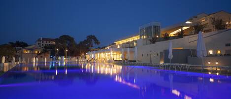 A nighttime view of a swimming pool with a blue-lit edge, surrounded by buildings with warm lighting.