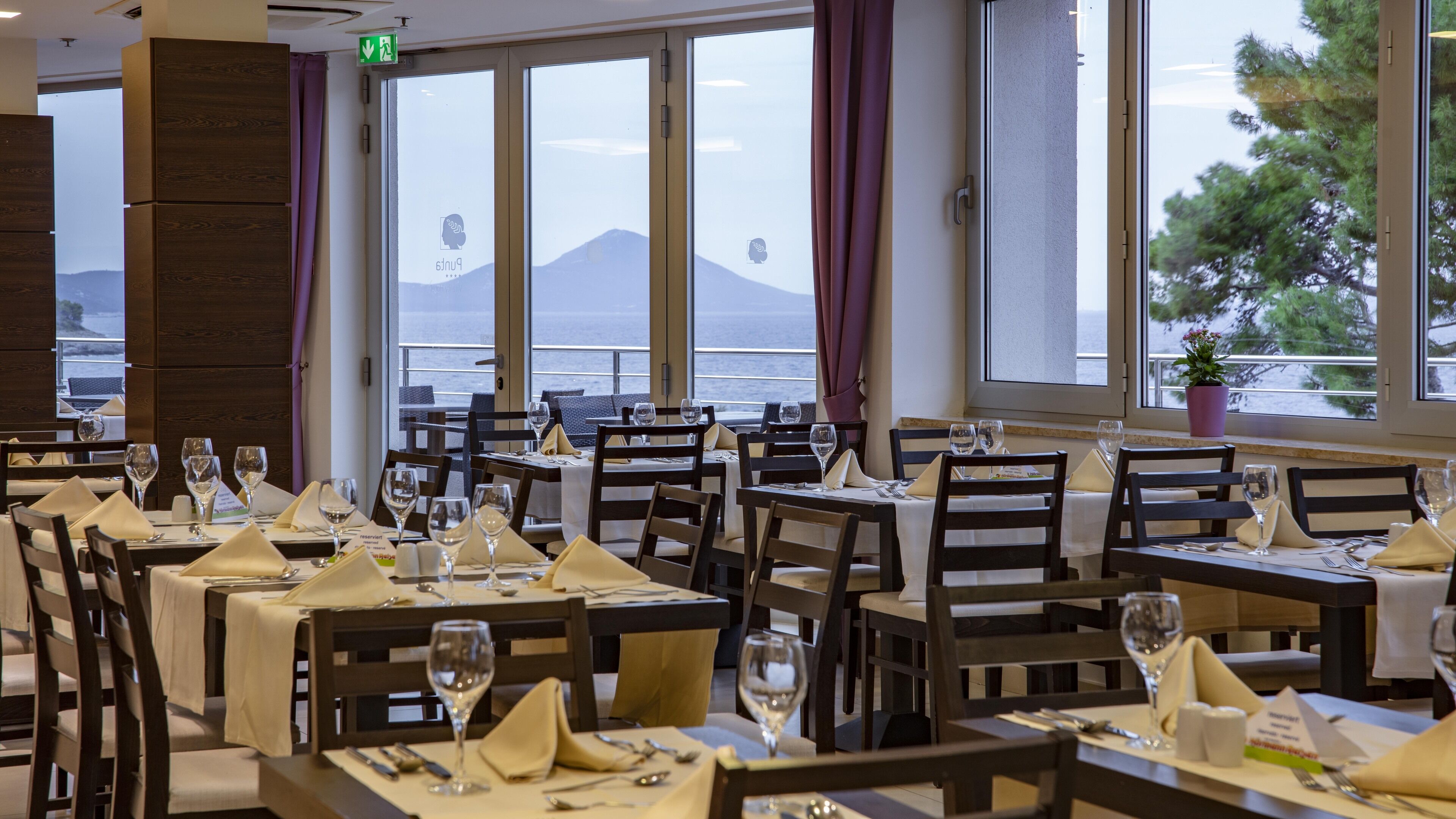 Ein Restaurant mit gedeckten Tischen, großen Fenstern mit Blick auf Meer und Berge sowie einem Blumentopf auf dem Fensterbrett.
