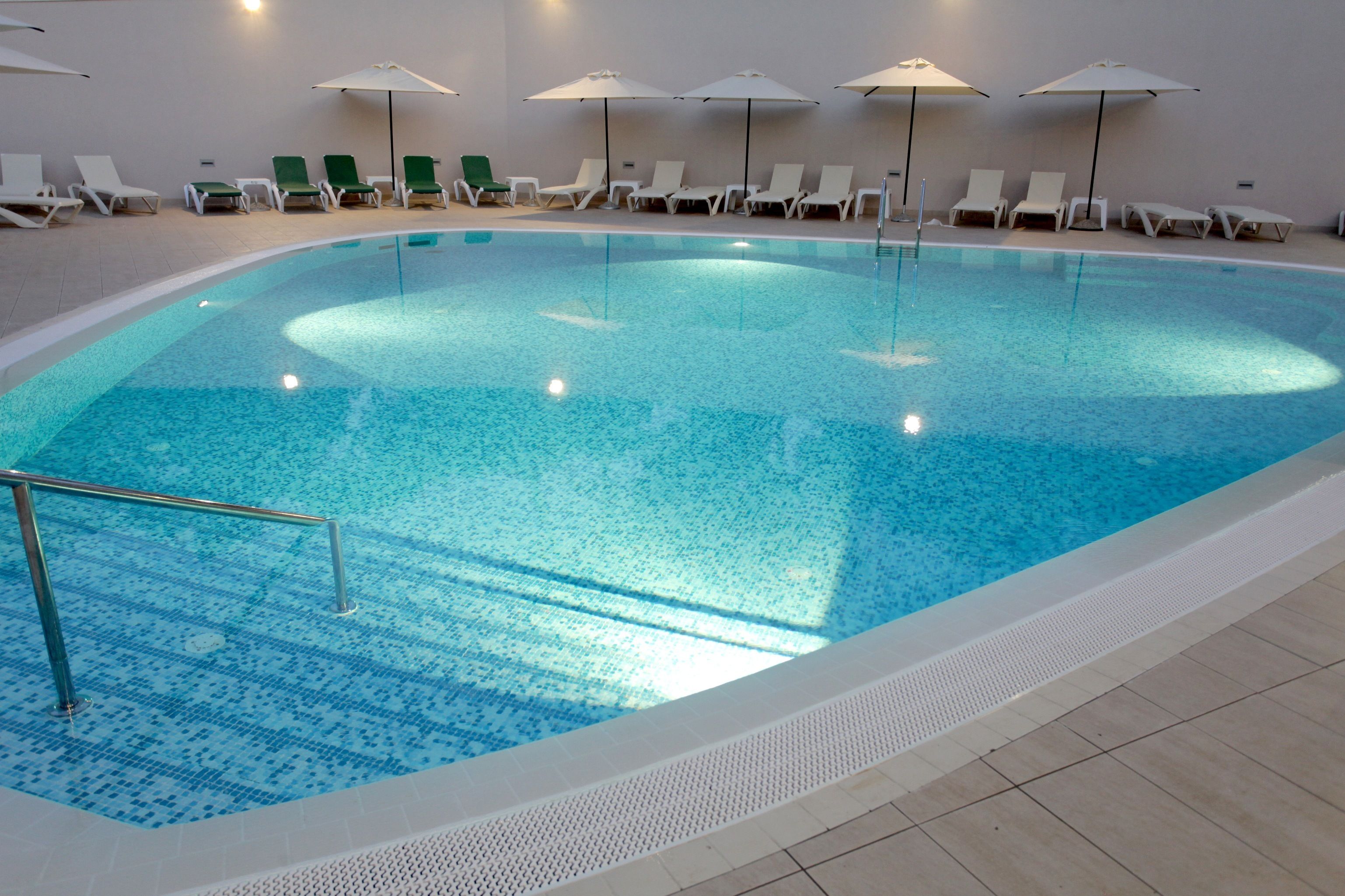En swimmingpool med flisebelagte omgivelser, omgivet af liggestole og parasoller.