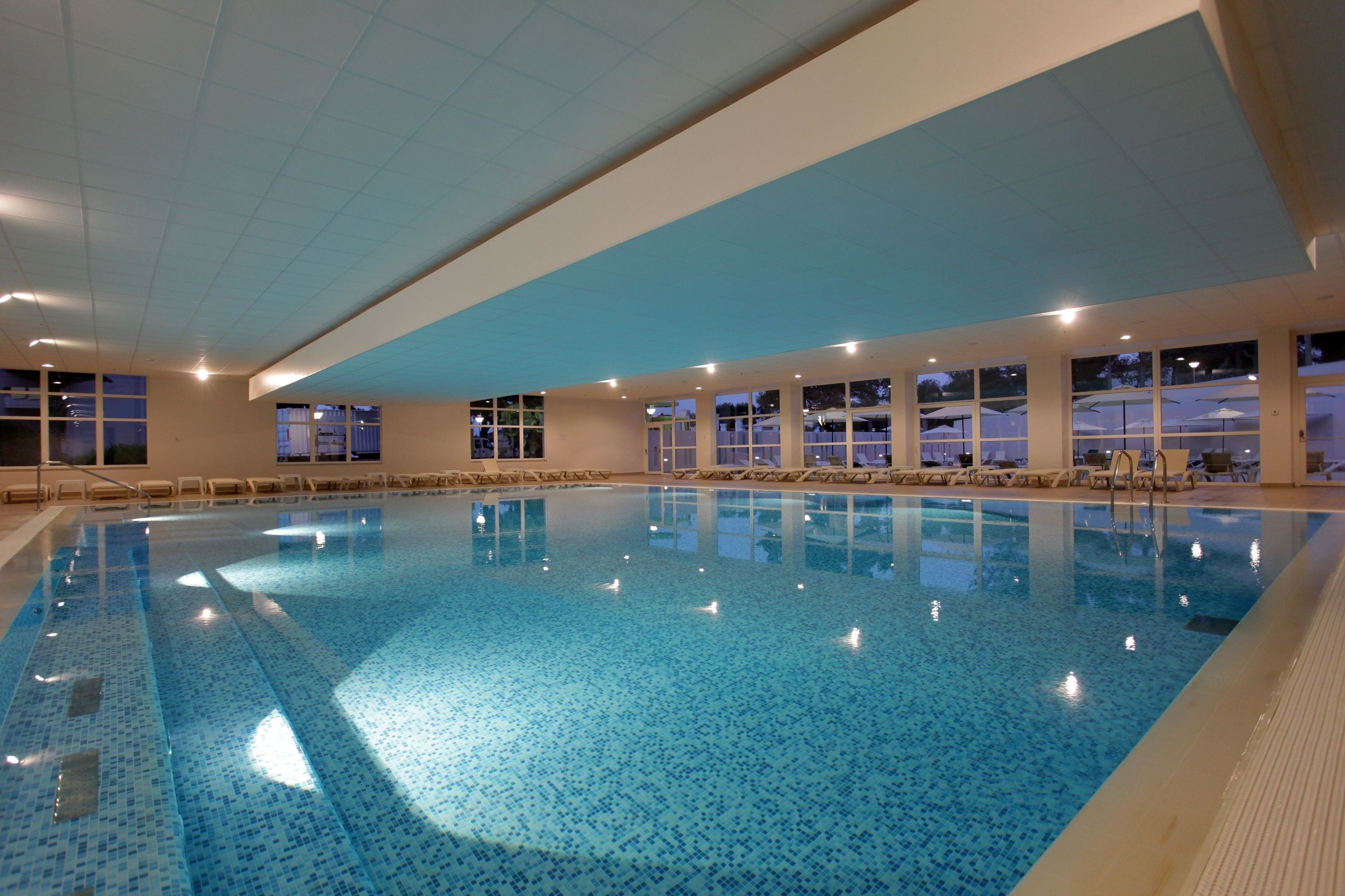 An indoor swimming pool with clear water, surrounded by windows and lounge chairs.