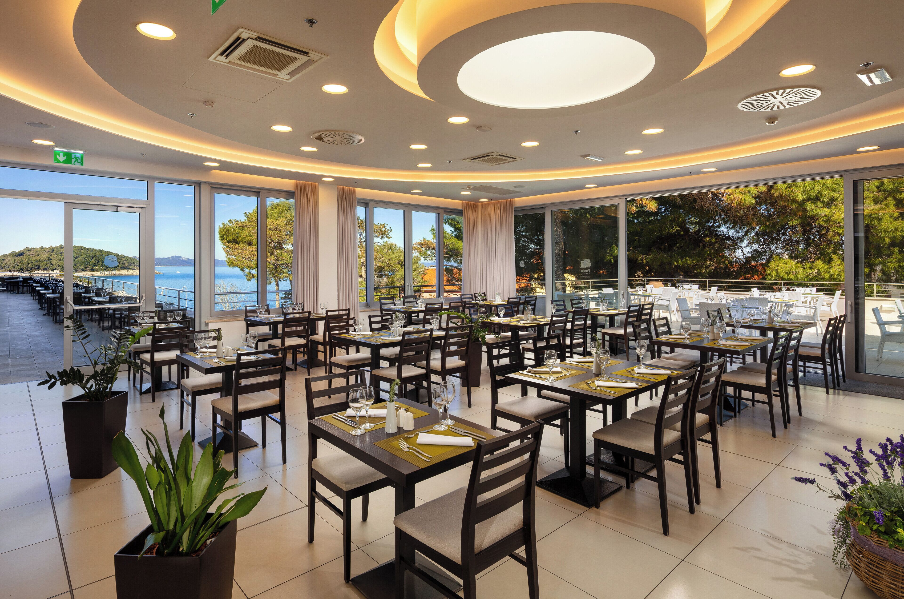 A spacious restaurant with multiple tables and chairs, large windows offering a view of the sea, and a modern ceiling design.