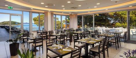 A spacious restaurant with multiple tables and chairs, large windows offering a view of the sea, and a modern ceiling design.