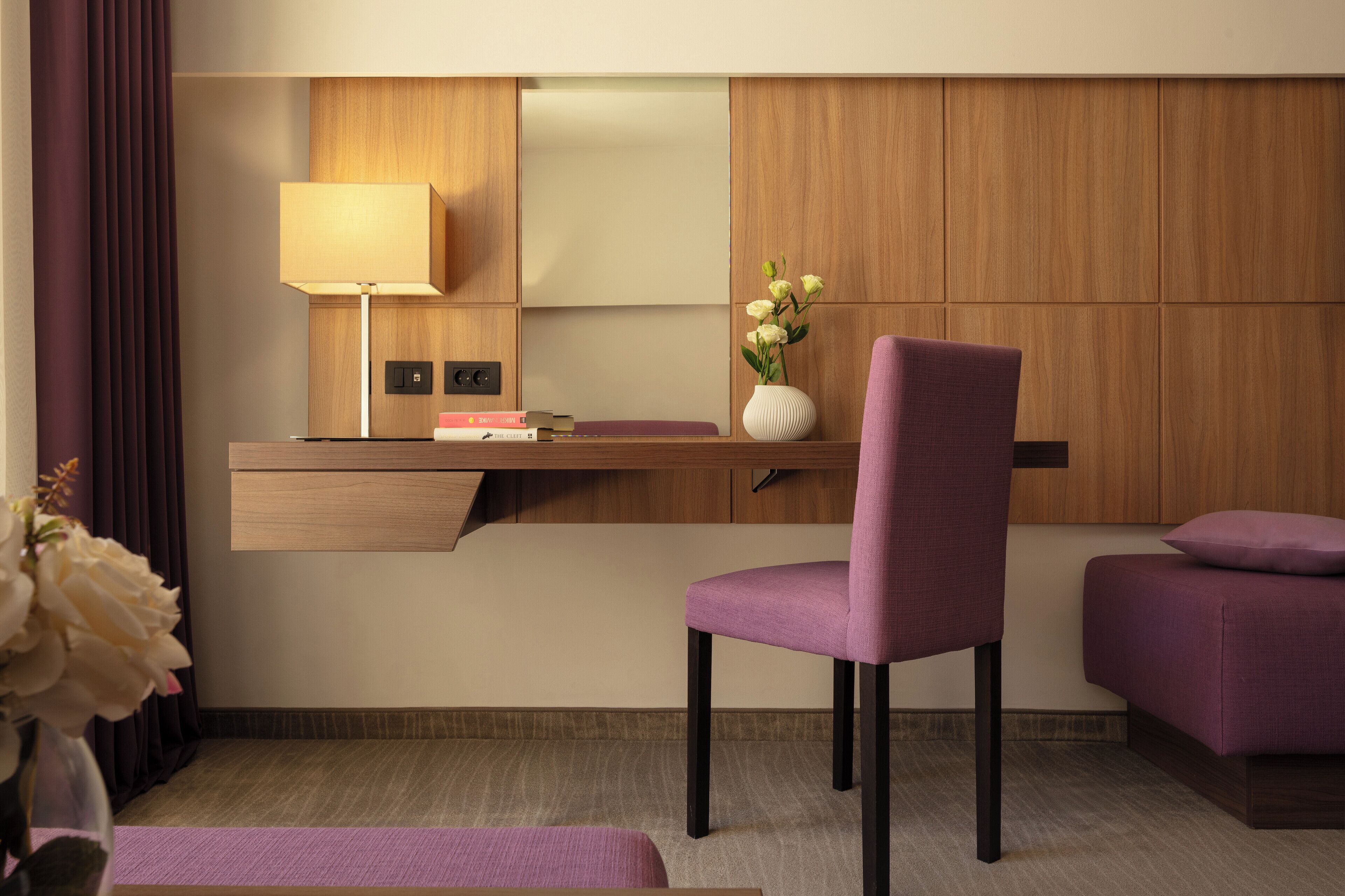 A modern hotel room with a wooden desk, a purple chair, a small sofa, a lamp, and a vase with flowers.