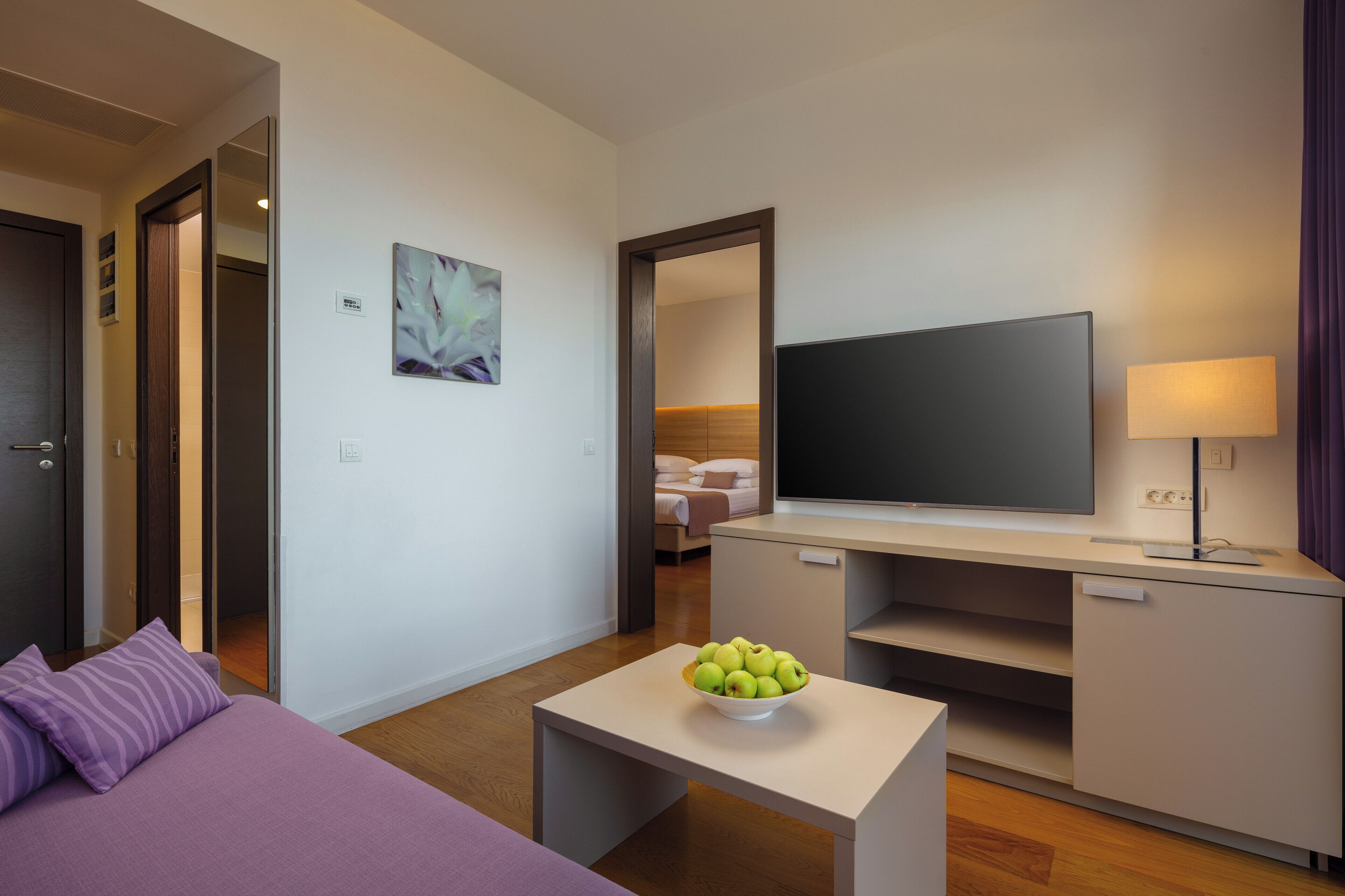 A hotel room with a bed, a small table with a bowl of fruit, a TV, a lamp, and a mirror.