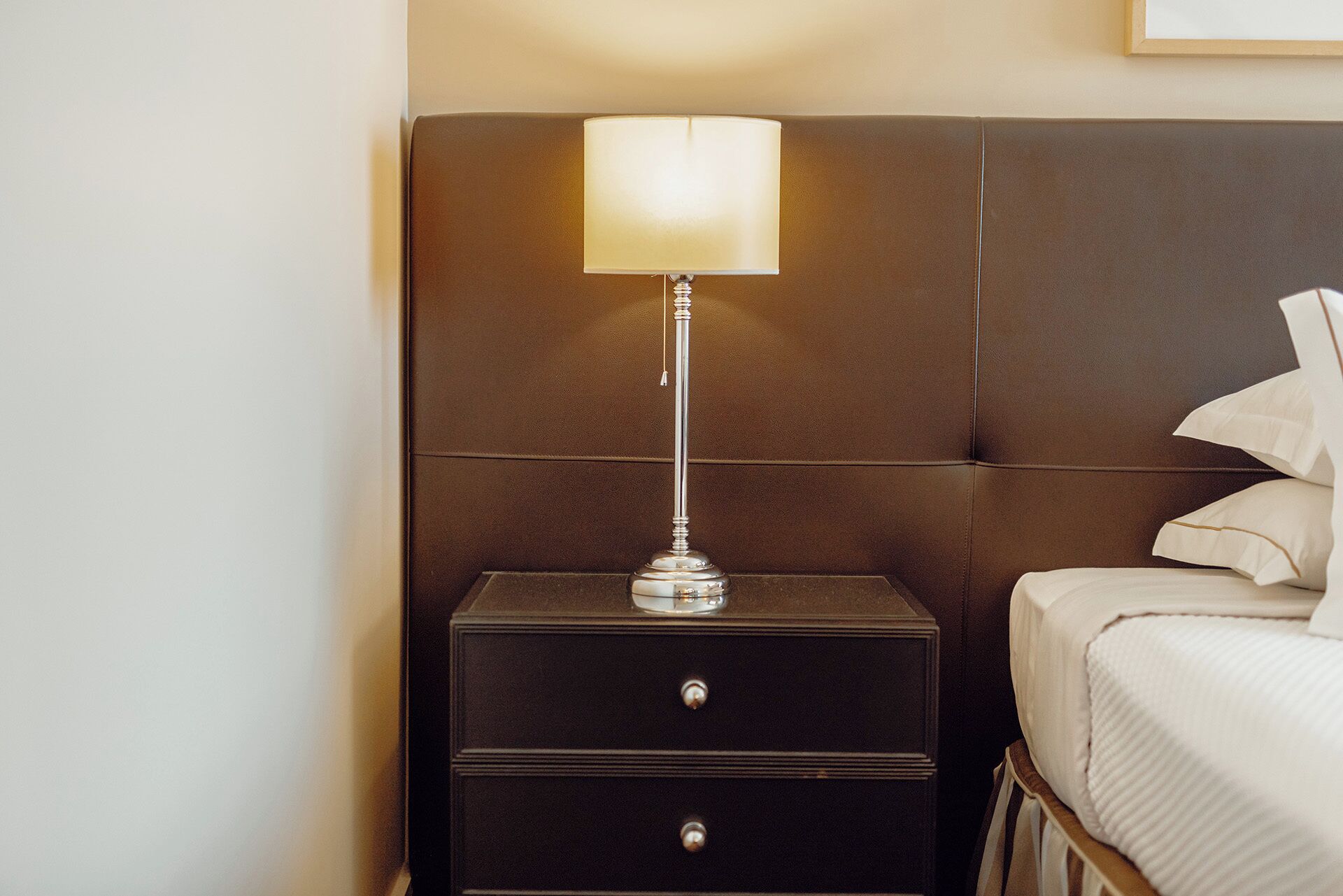 A neatly made bed with white linens, a bedside table with a lamp, and a dark headboard.
