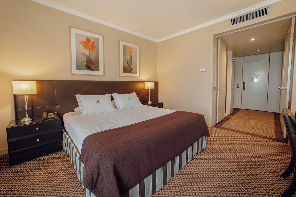 A hotel room with a large bed, two bedside lamps, a nightstand, a dresser, and a door leading to another room.
