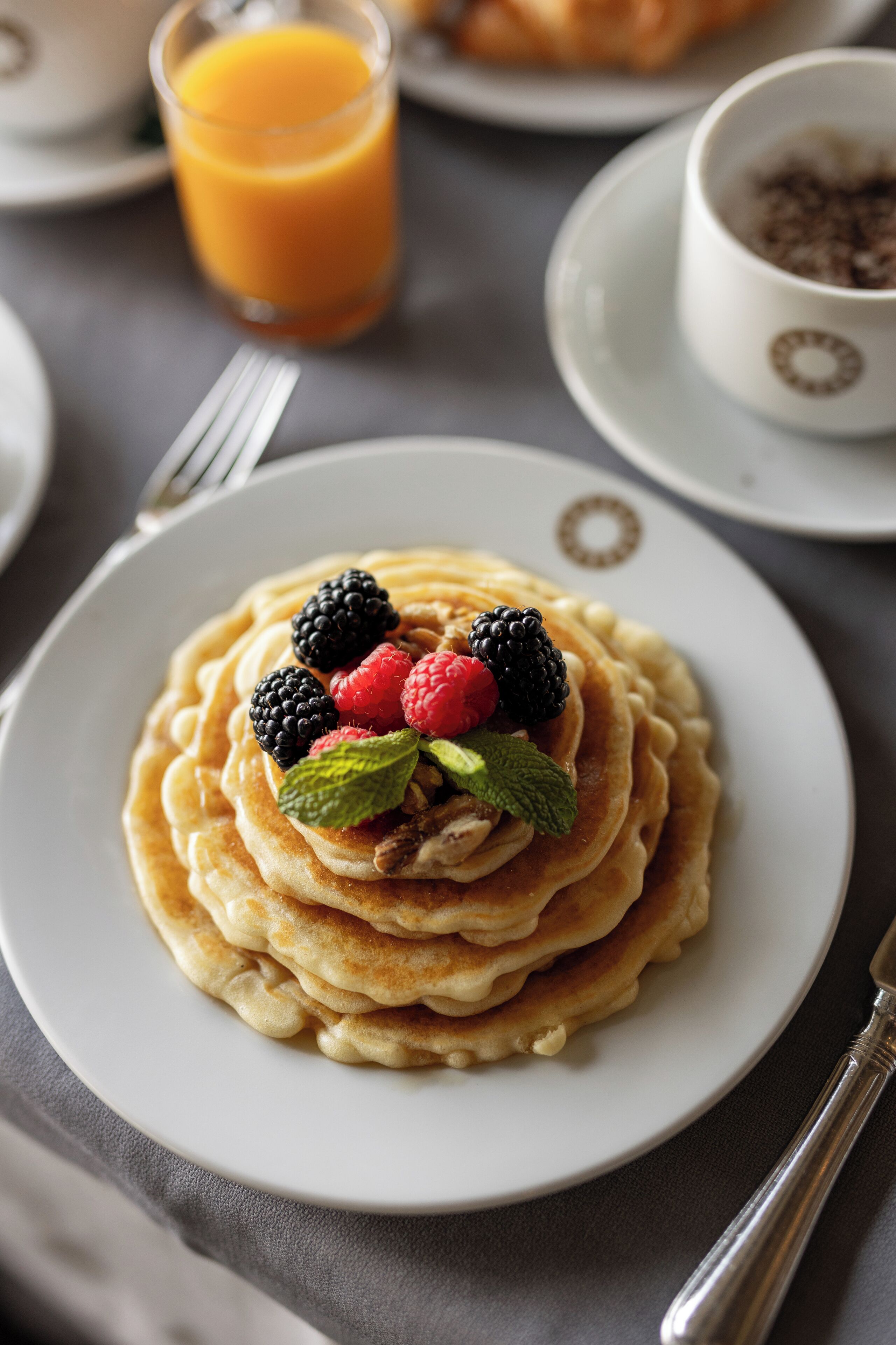 Un piatto di pancake con frutti di bosco e menta su un tavolo, insieme a un bicchiere di succo d'arancia e una tazza.