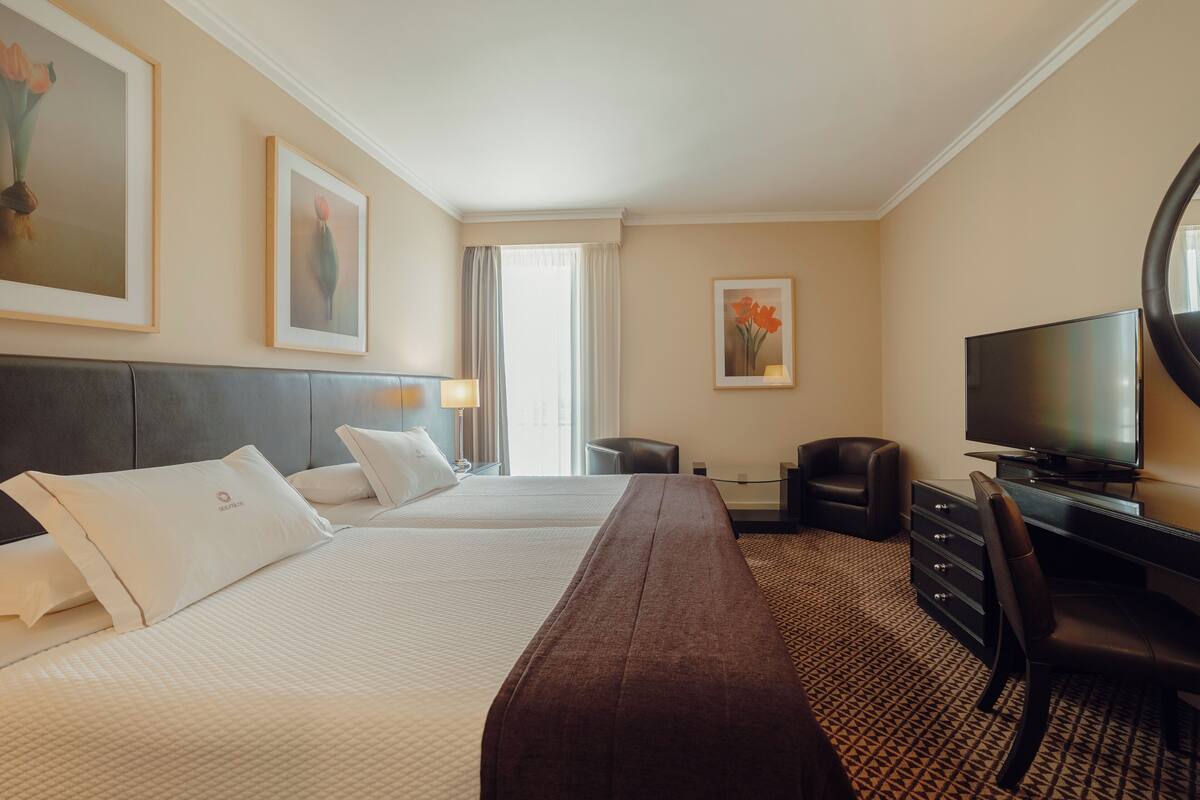 A hotel room with two beds, a desk, a chair, a TV, and artwork on the walls.