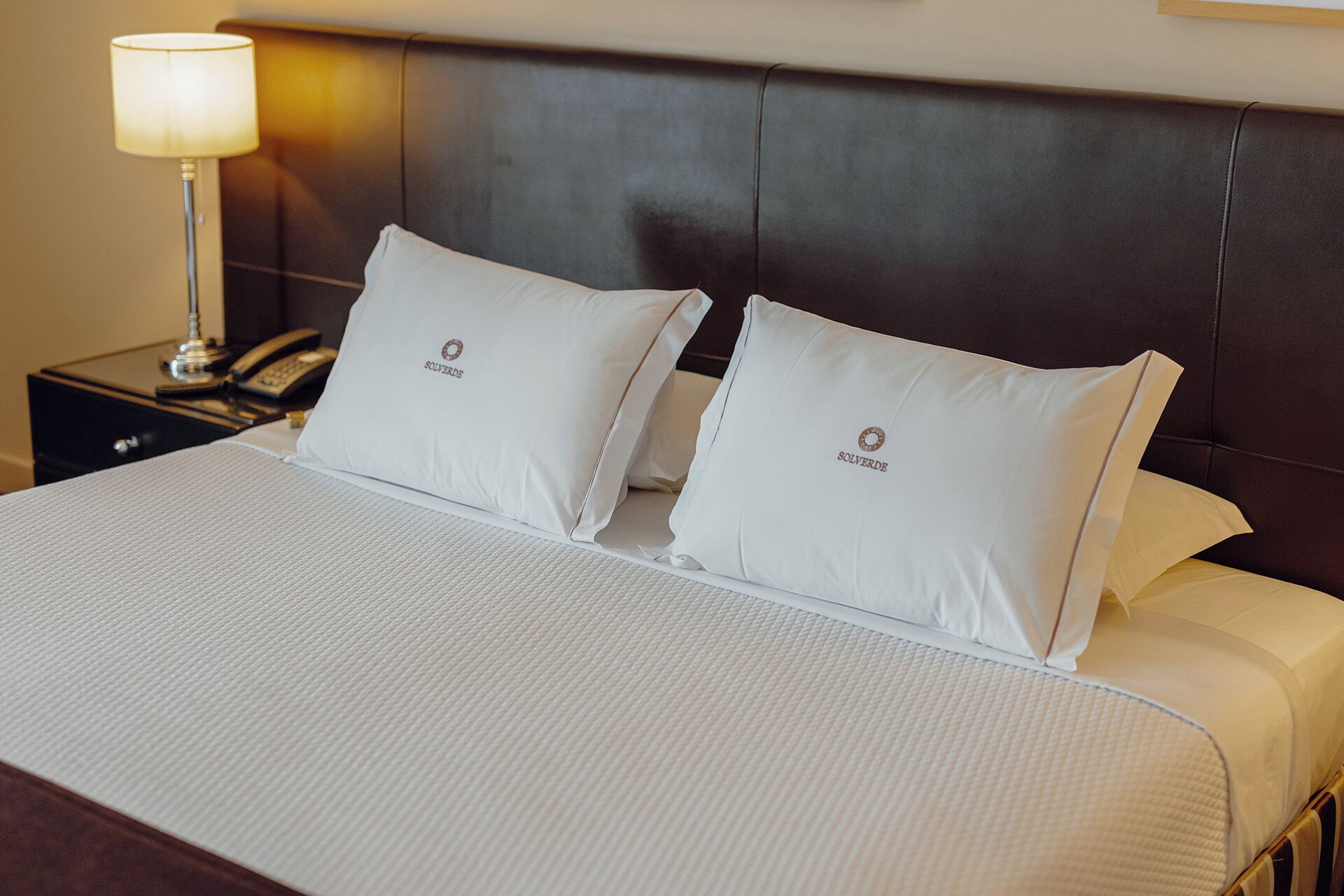 Habitación de hotel con una cama grande, dos almohadas con un logotipo, una lámpara de noche y una mesita de noche con un teléfono.