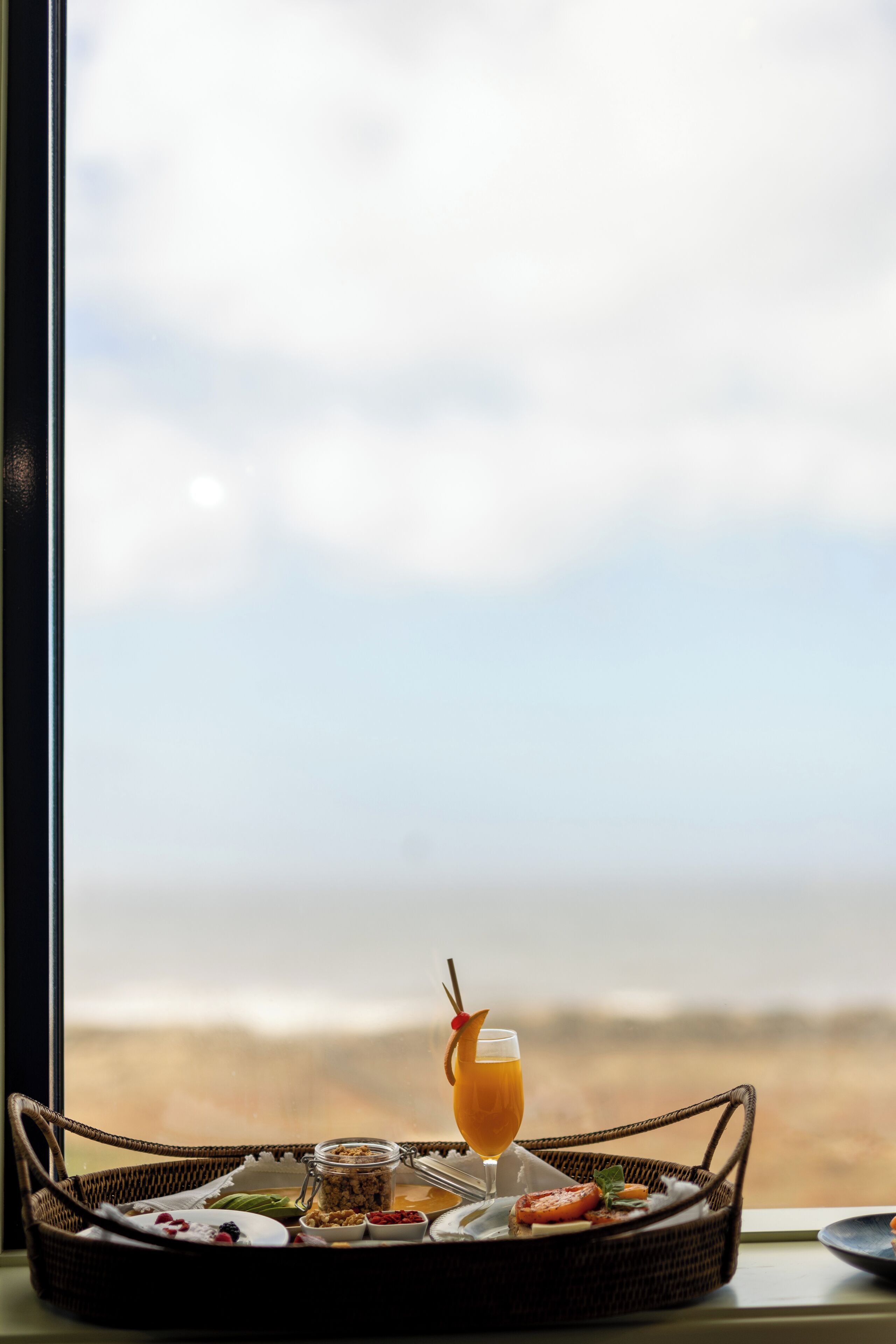 Un plateau de petit-déjeuner avec un verre de jus d’orange, un pot de confiture et diverses pâtisseries, posé sur une table en osier près d’une fenêtre donnant sur la plage.