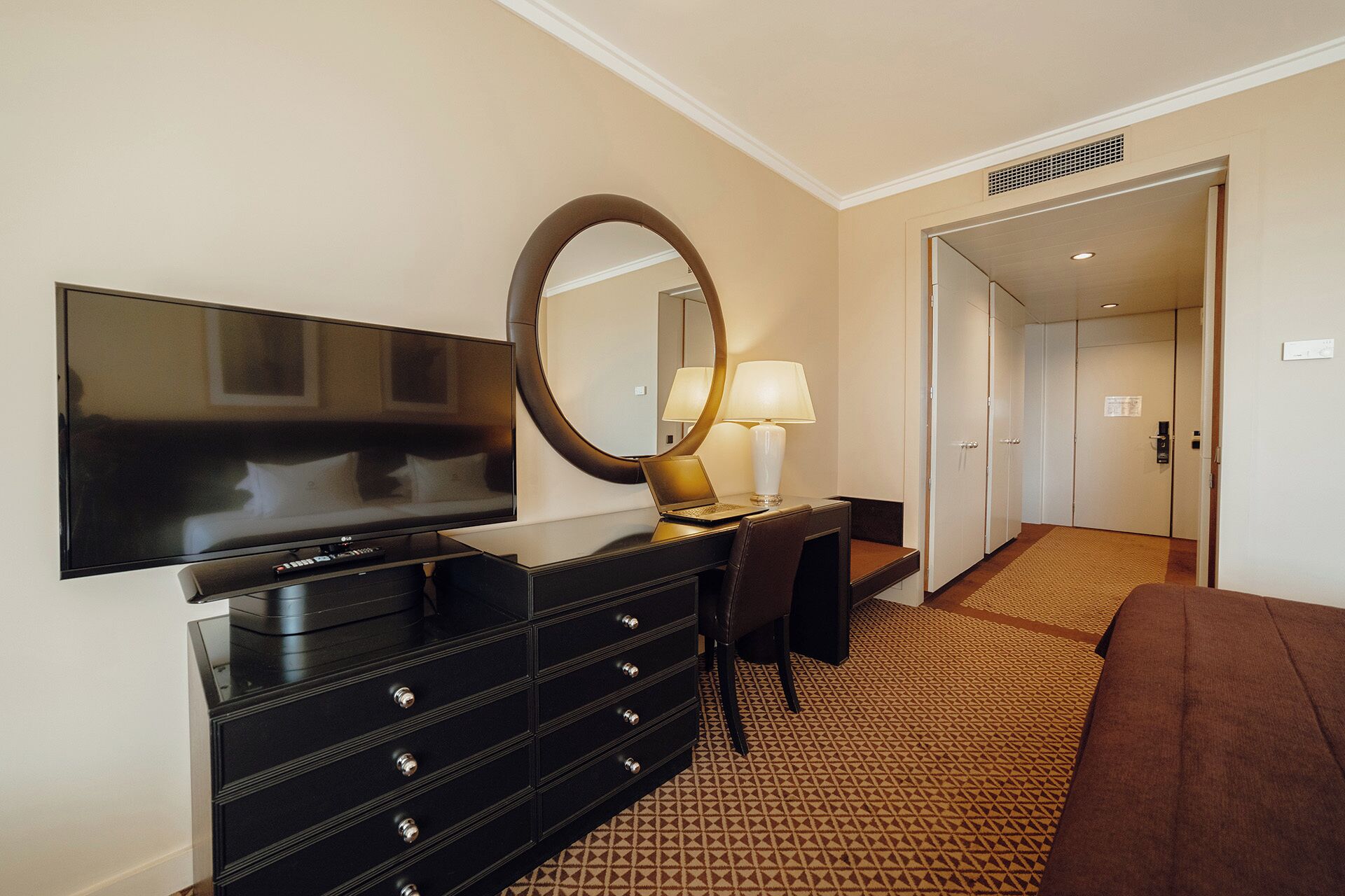 Habitación de hotel con televisión de pantalla plana, escritorio con lámpara, espejo grande, cómoda y alfombra estampada.