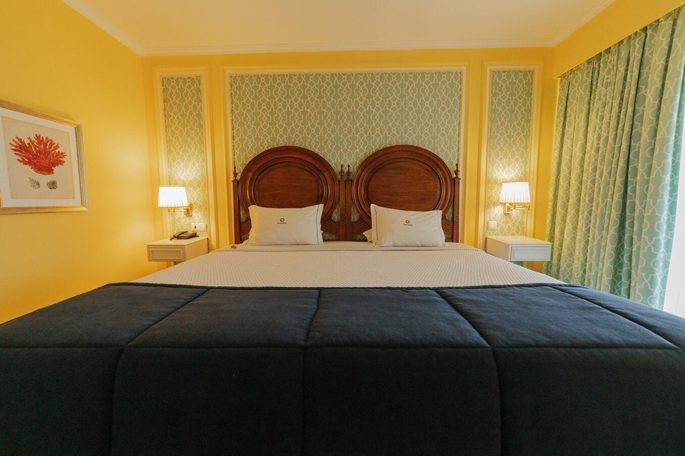 Habitación de hotel con una cama grande, dos mesitas de noche, un cuadro enmarcado en la pared y papel tapiz estampado.