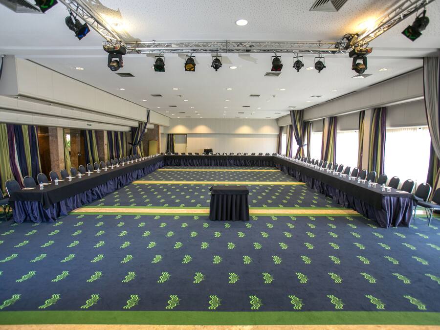 Un salón de conferencias con una larga mesa, filas de sillas y un escenario con equipo de iluminación.