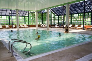 An indoor swimming pool with clear water, surrounded by lounge chairs and a covered area with large windows.