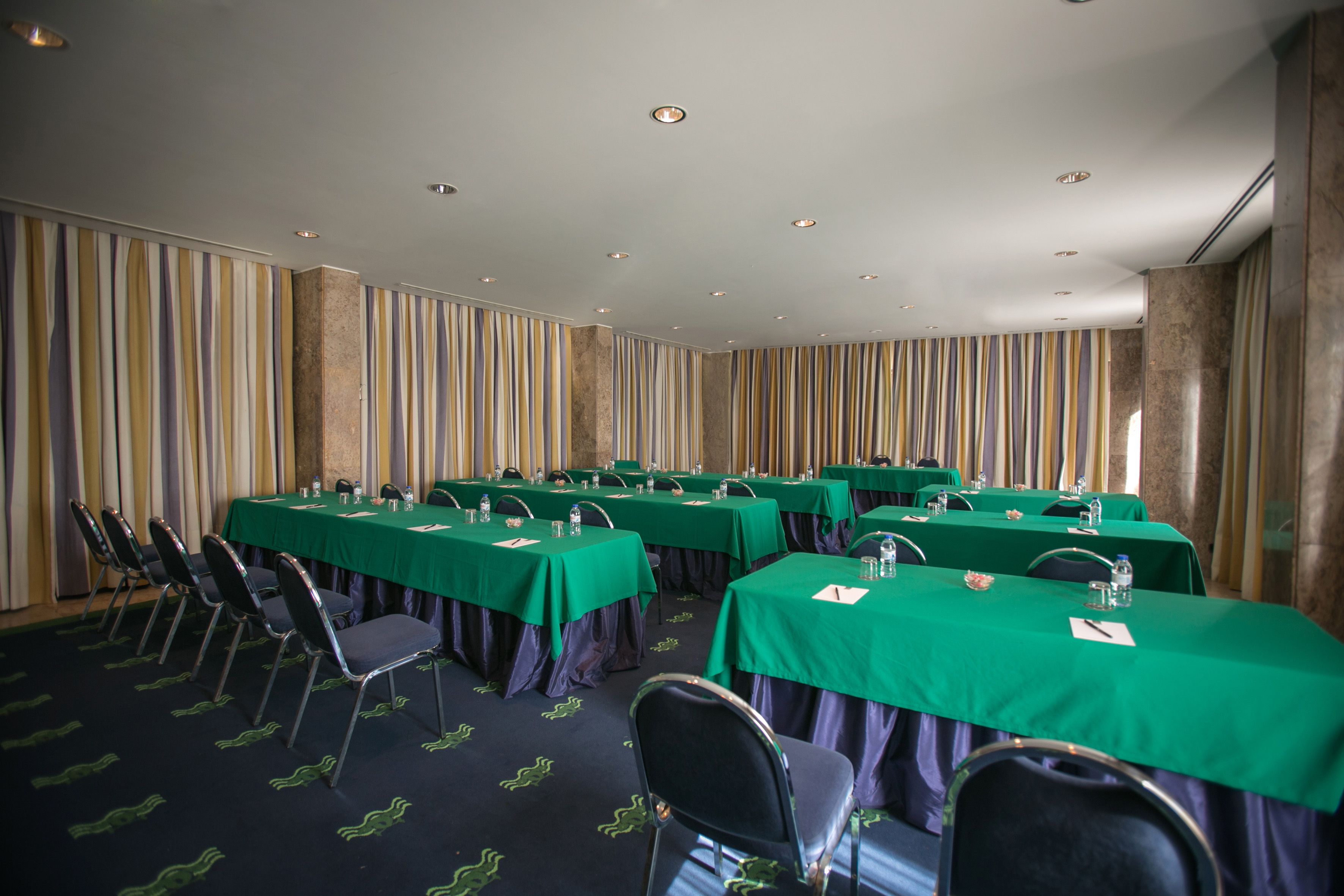 Un salón de conferencias con varias mesas y sillas, manteles verdes y un telón a rayas como fondo.