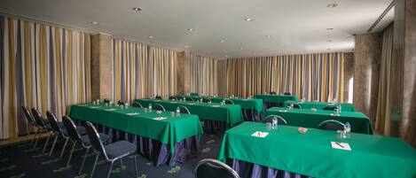 Un salón de conferencias con varias mesas y sillas, manteles verdes y un telón a rayas como fondo.