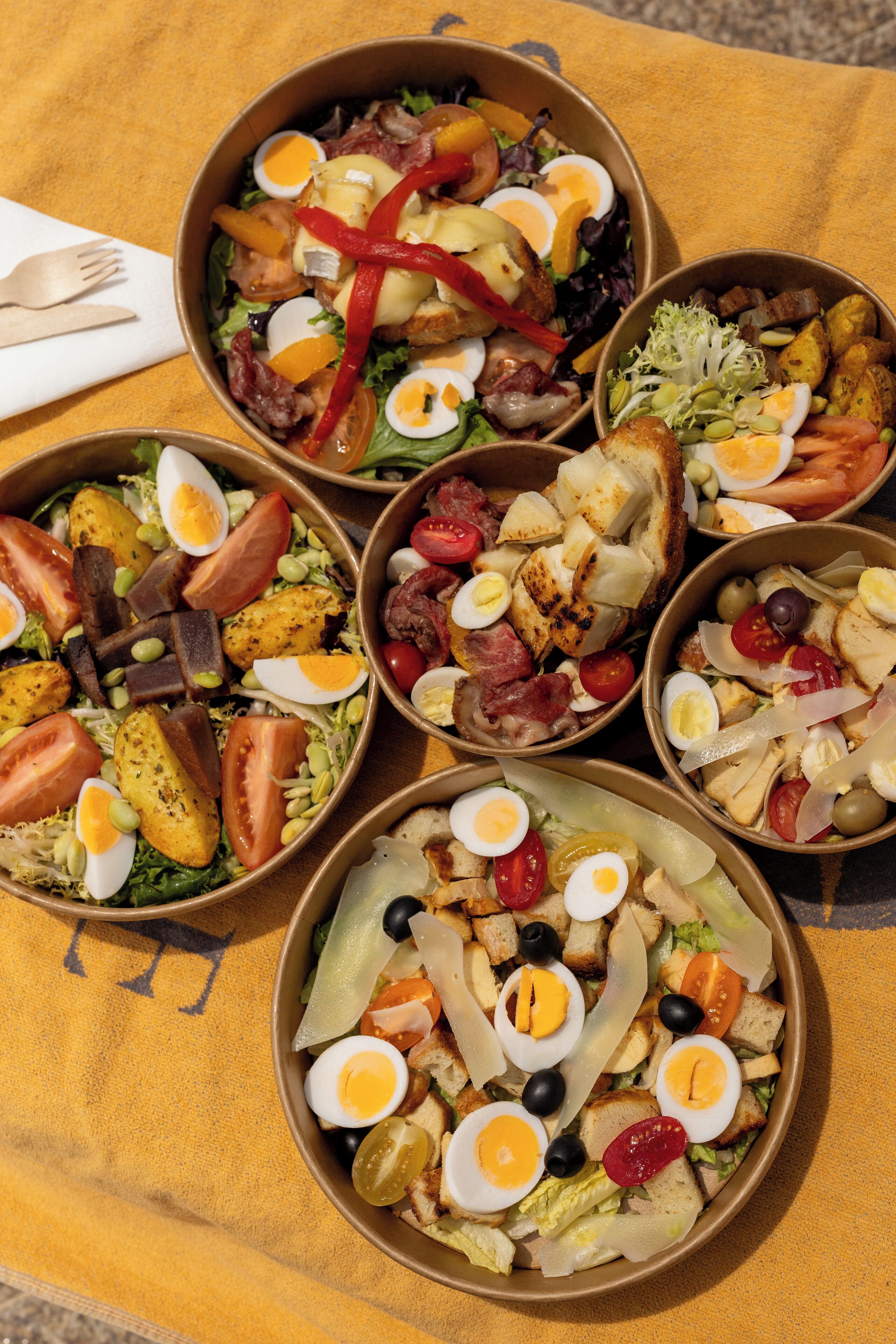 A table with multiple bowls of salad containing various ingredients like tomatoes, eggs, and greens.