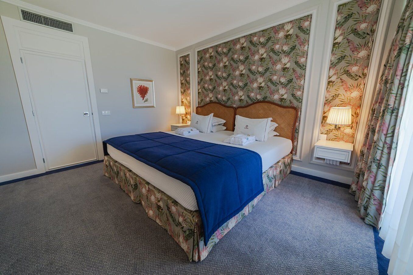 Habitación de hotel con una cama grande, papel tapiz floral, cabecera, mesita de noche con lámpara y una puerta.