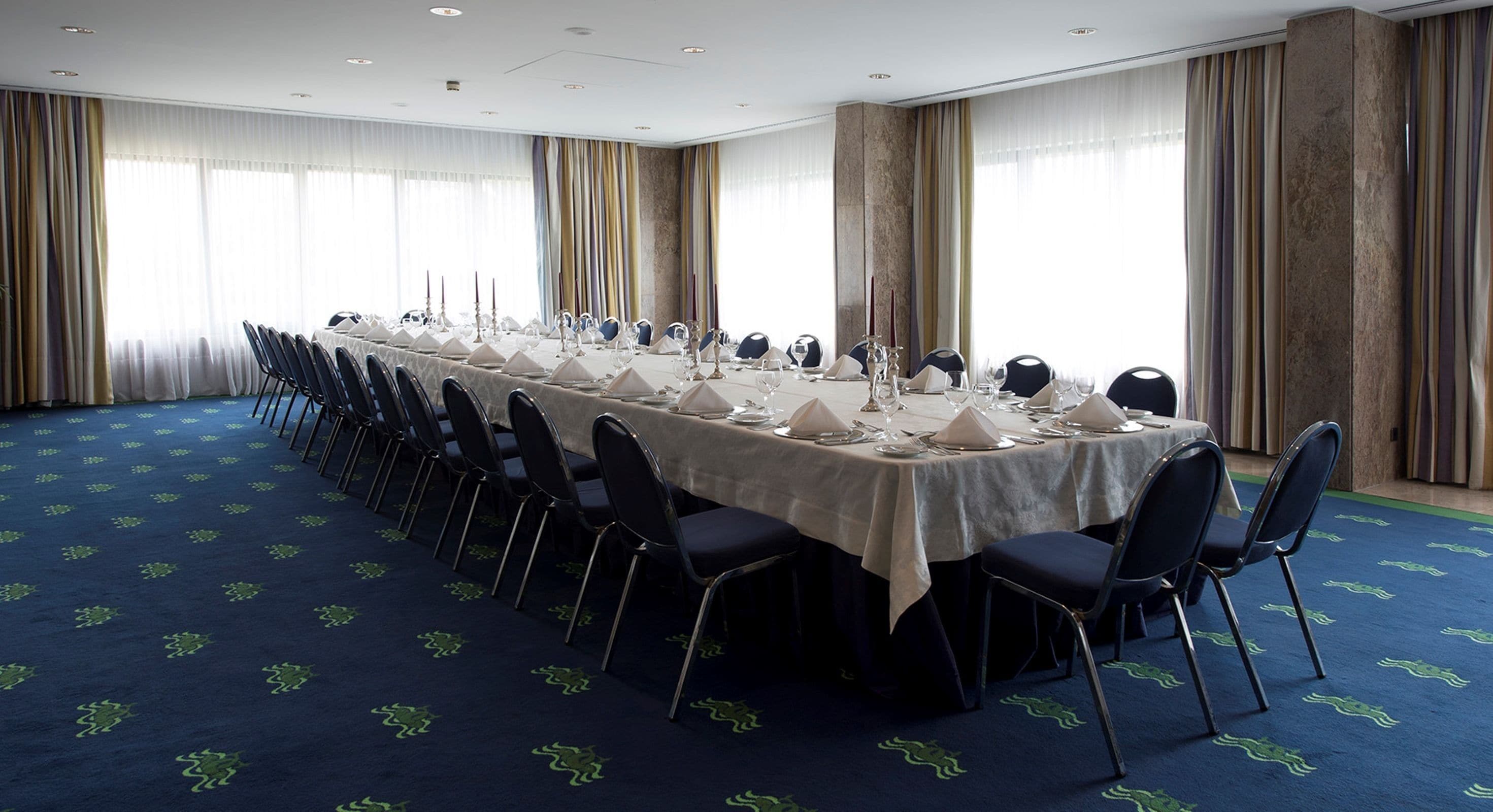 A conference room with a long table set for a meeting, surrounded by chairs, with a carpeted floor and large windows with curtains.