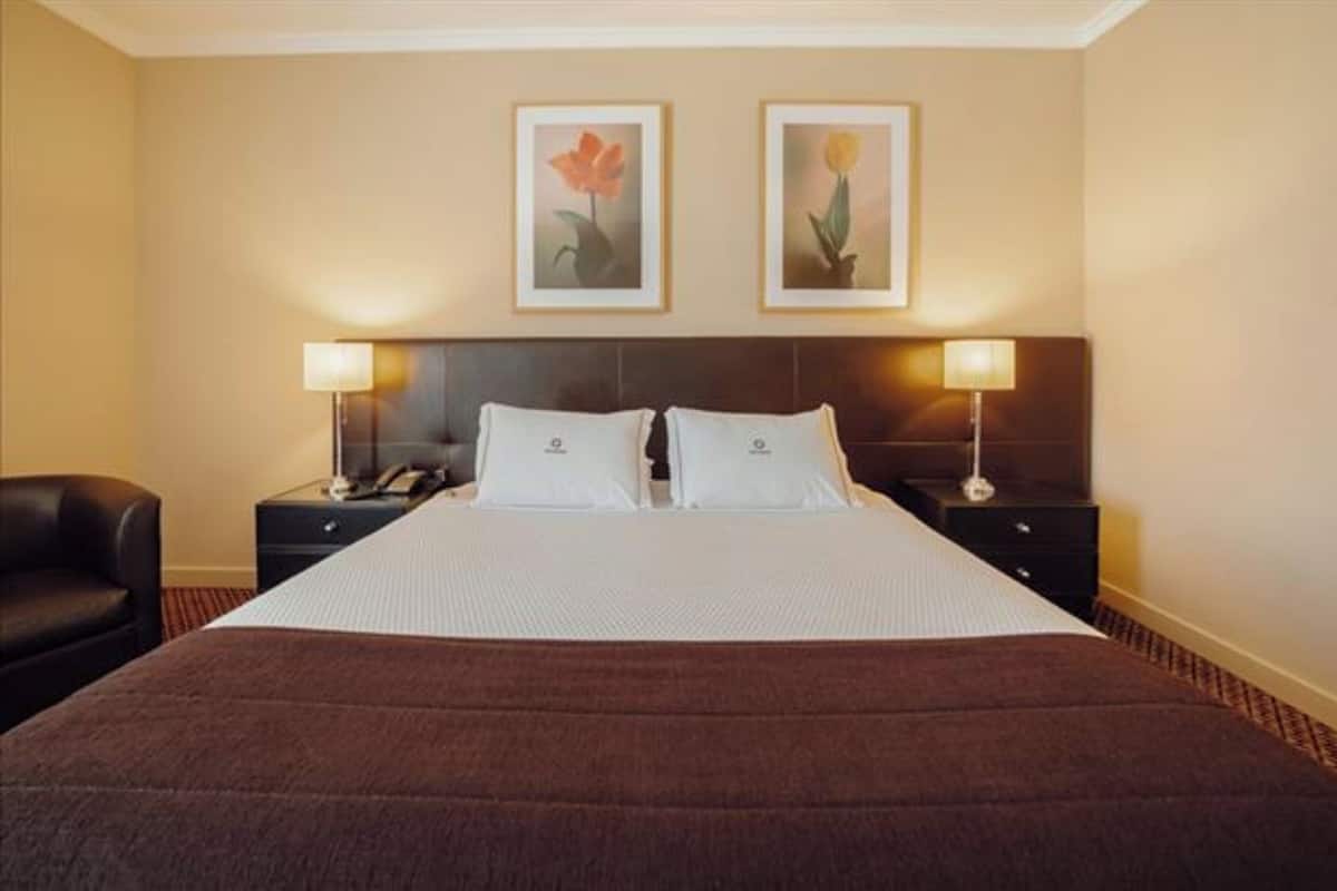 Habitación de hotel con una cama grande, dos mesitas de noche con lámparas, una silla y dos cuadros en la pared.
