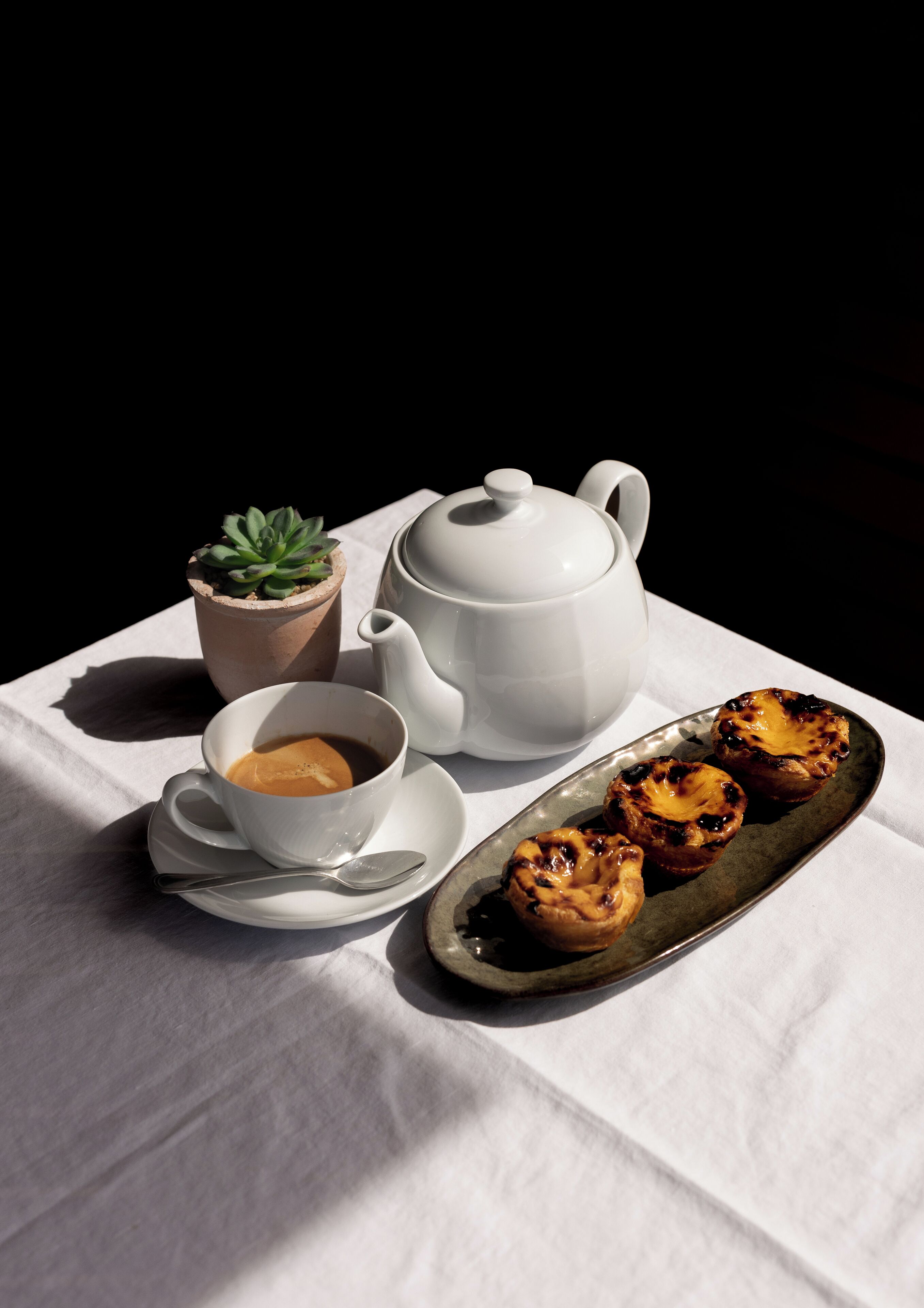 A table with a white tablecloth, a white teapot, a cup of coffee, a small potted plant, and a plate of pastries.
