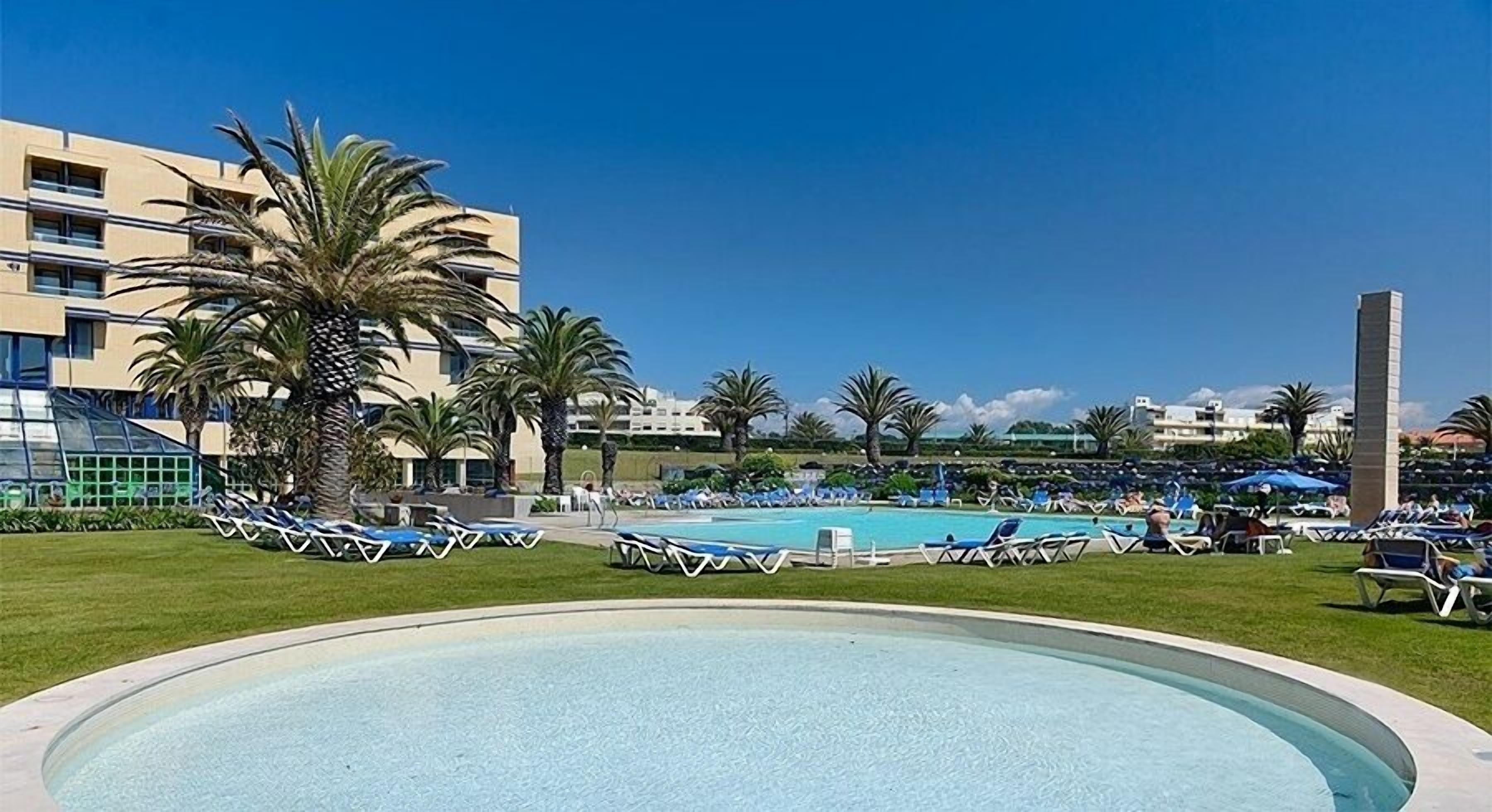 Zona de piscina con sillas de descanso y palmeras, rodeada de edificios.