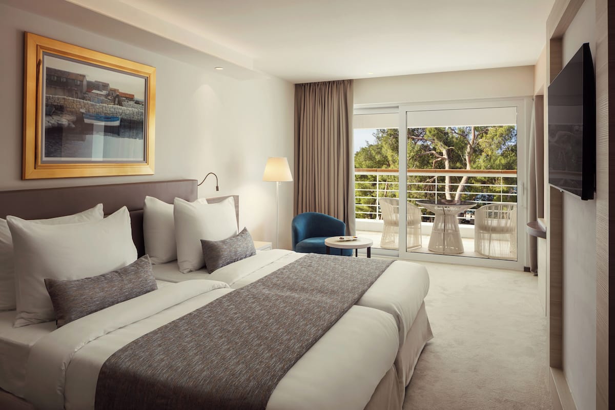 Ein Hotelzimmer mit einem großen Bett, einem Fernseher, einem gerahmten Bild, einem Sessel, einem kleinen Tisch und einem Balkon mit Blick auf Bäume.
