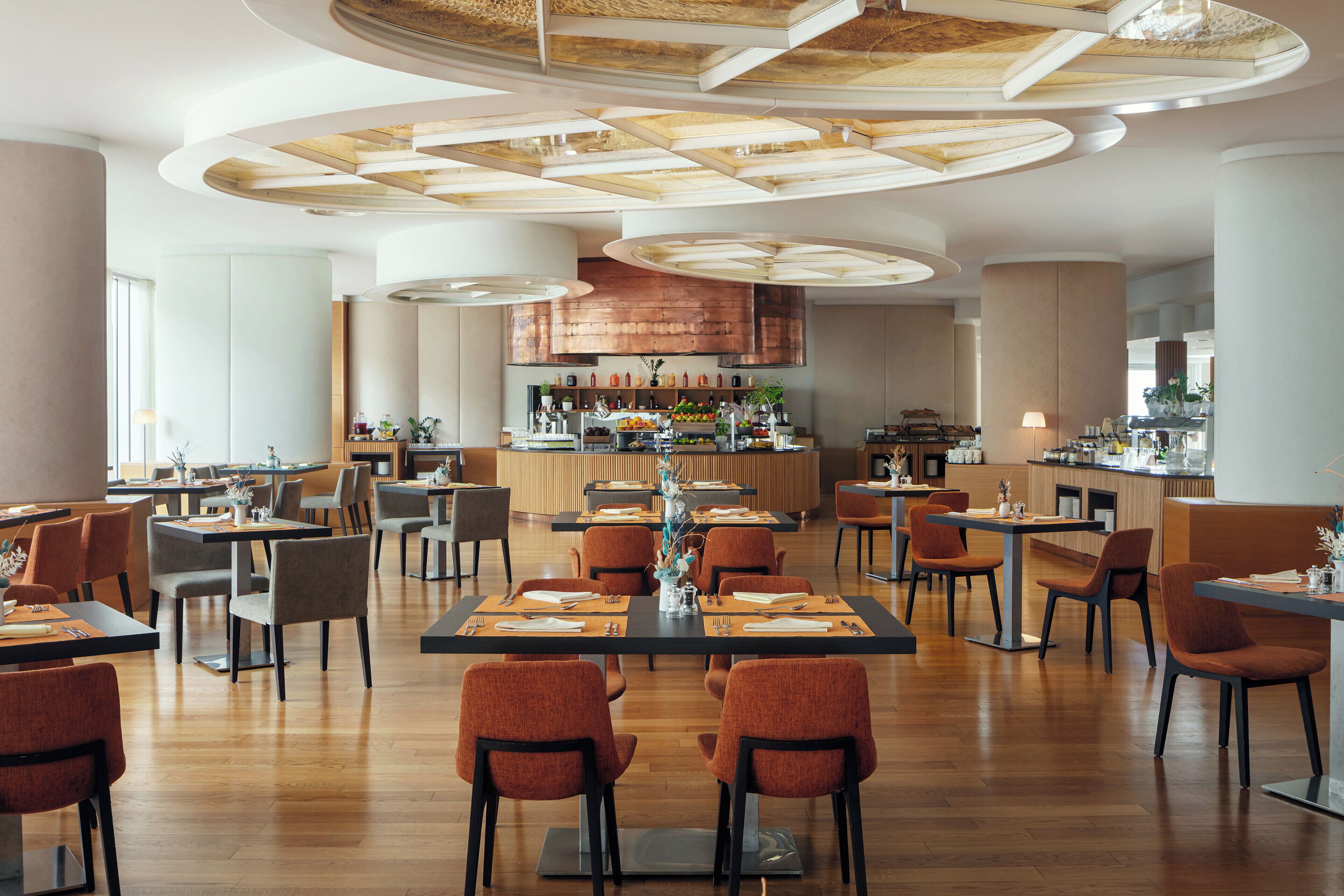 A modern restaurant with wooden flooring, round tables, and orange chairs.