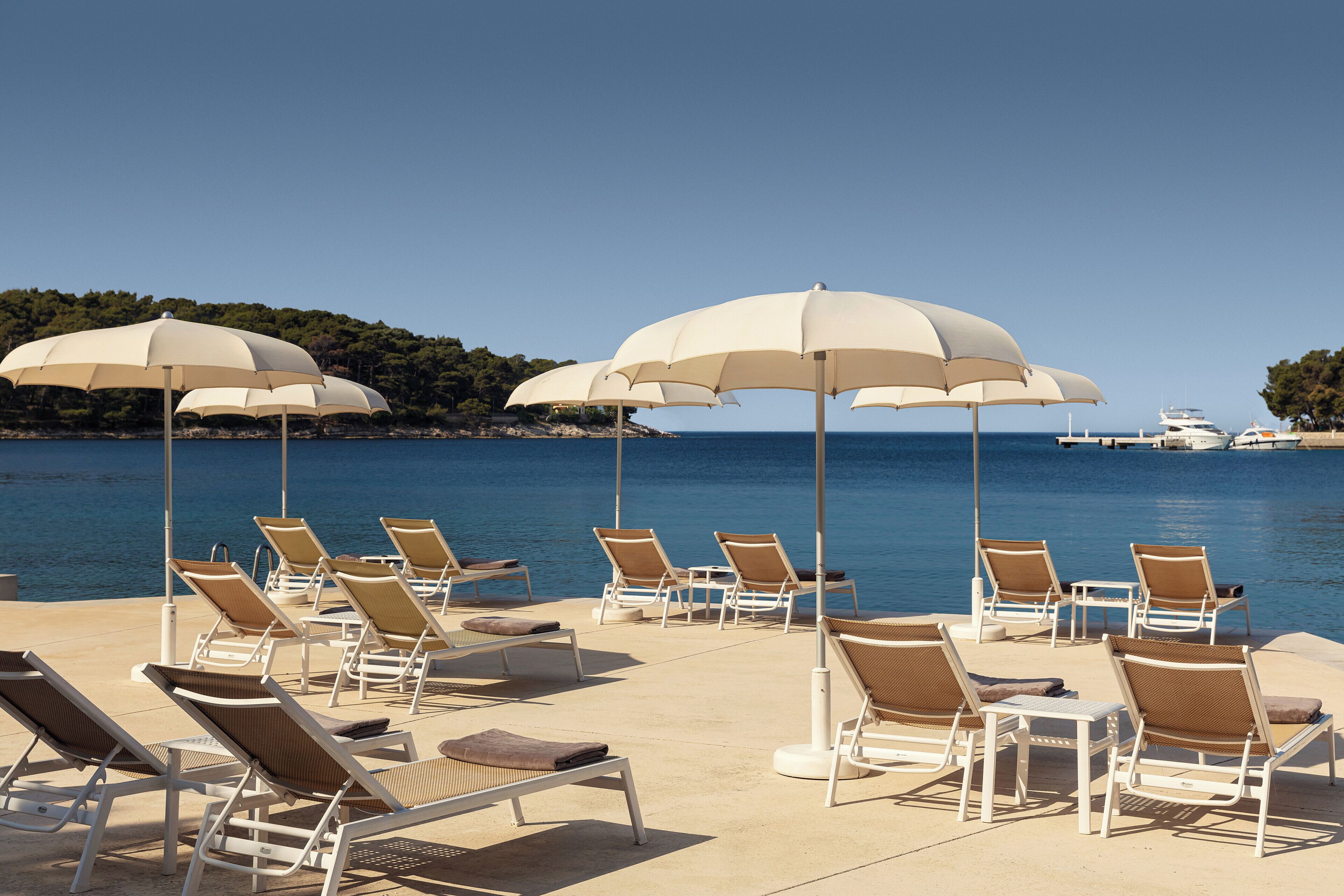 A beachside lounge area with sun loungers and umbrellas, overlooking a calm sea and a distant coastline.