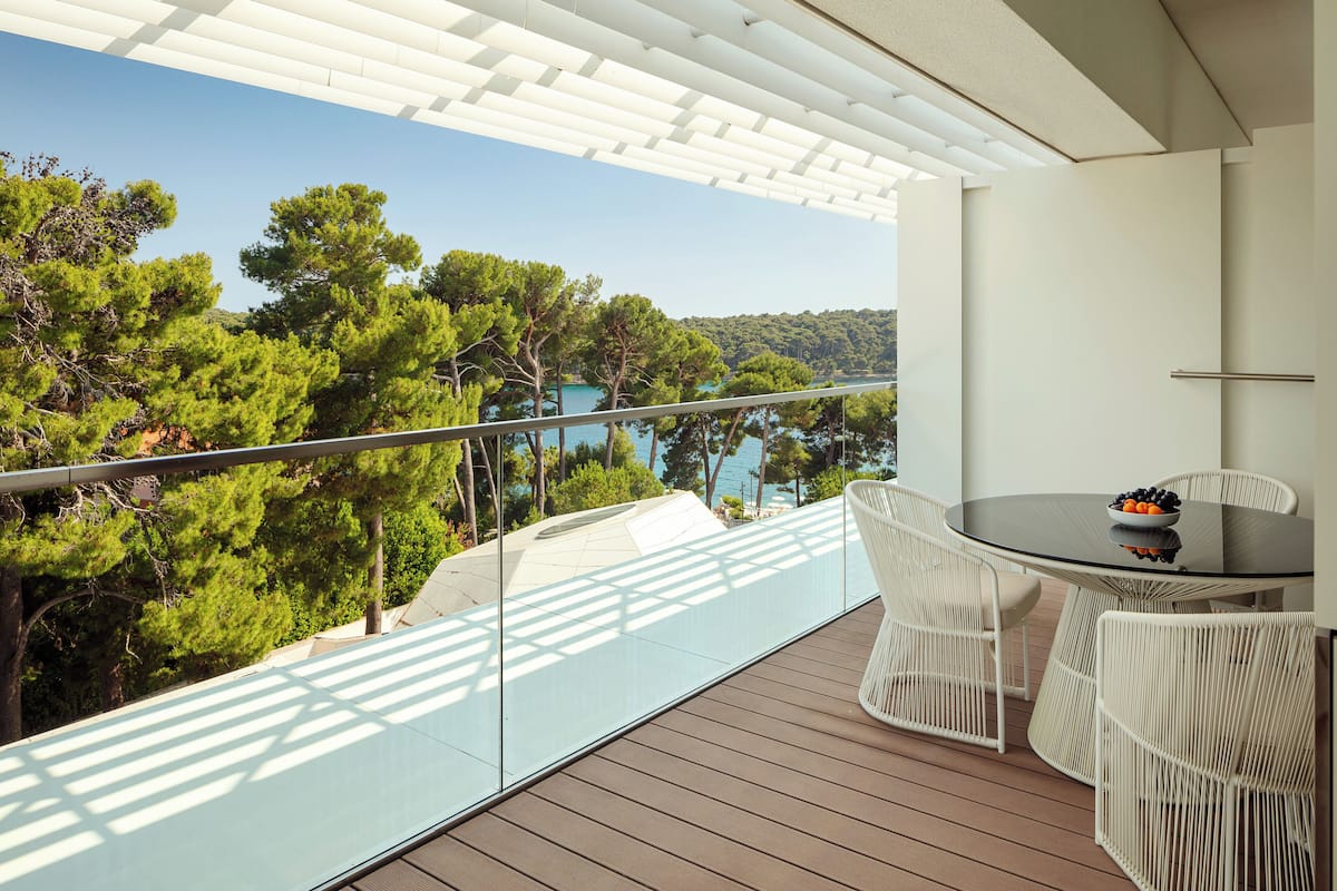 Ein Balkon mit Glastrennwand, weißen Korbstühlen, einem runden Tisch mit Obstschale und Blick auf Bäume und Wasser.