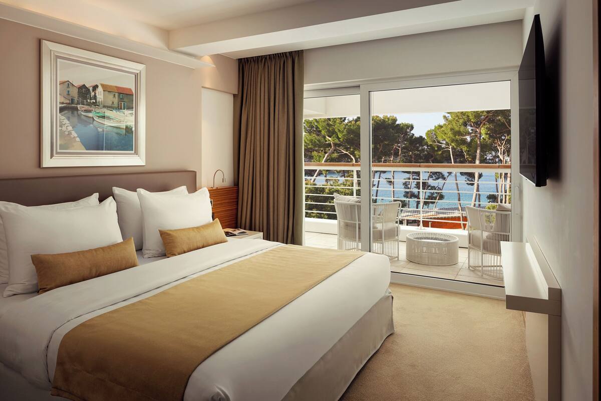 Ein Hotelzimmer mit einem großen Bett, Blick auf einen Pool und einem Balkon mit Sitzgelegenheit.