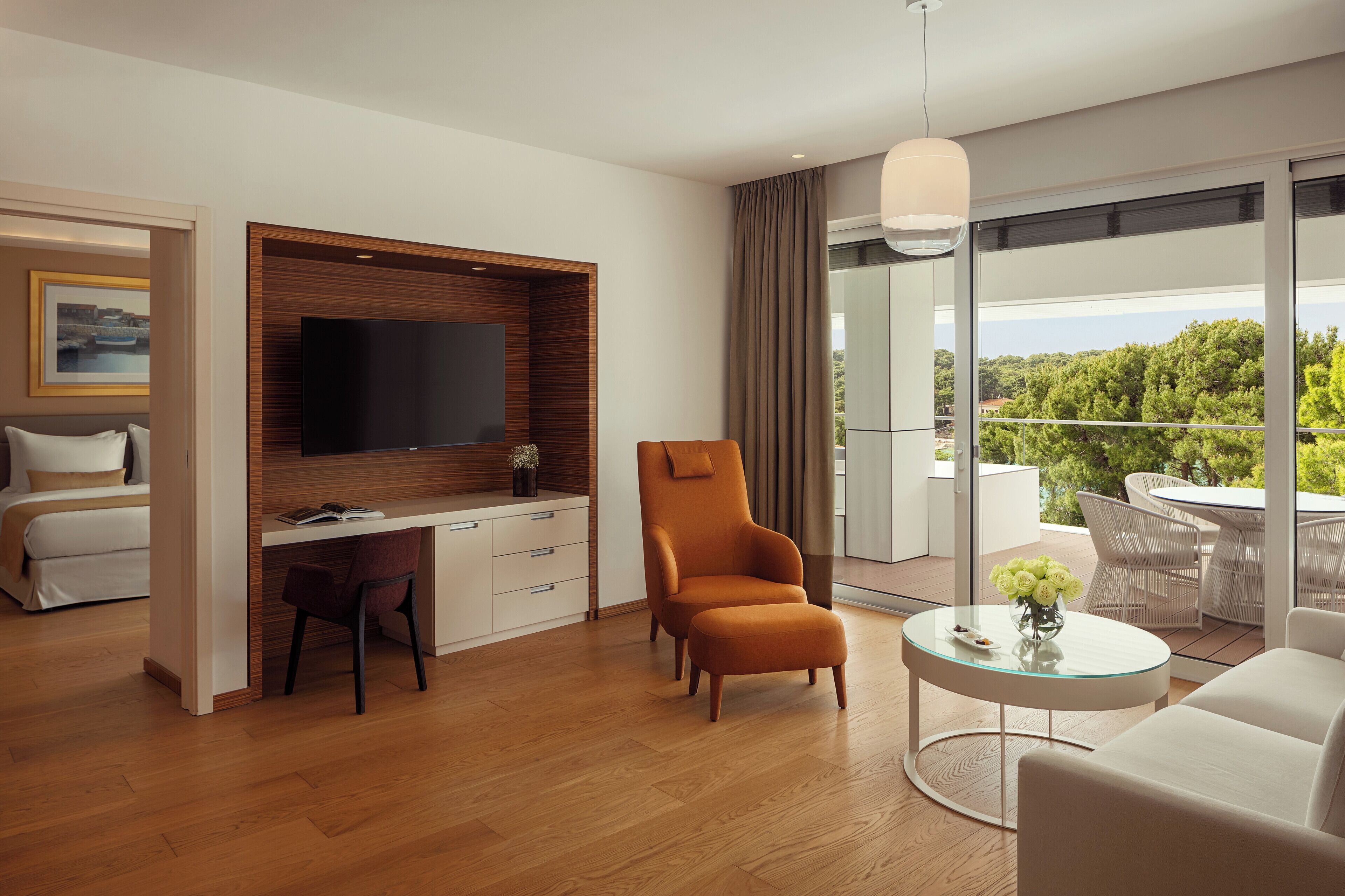 A modern hotel room with a wooden desk, a comfortable armchair, a round glass coffee table, and a balcony with outdoor furniture.
