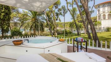 Un jacuzzi con un cuenco de madera, una mesita con toallas y botellas, y vistas a un edificio y árboles.