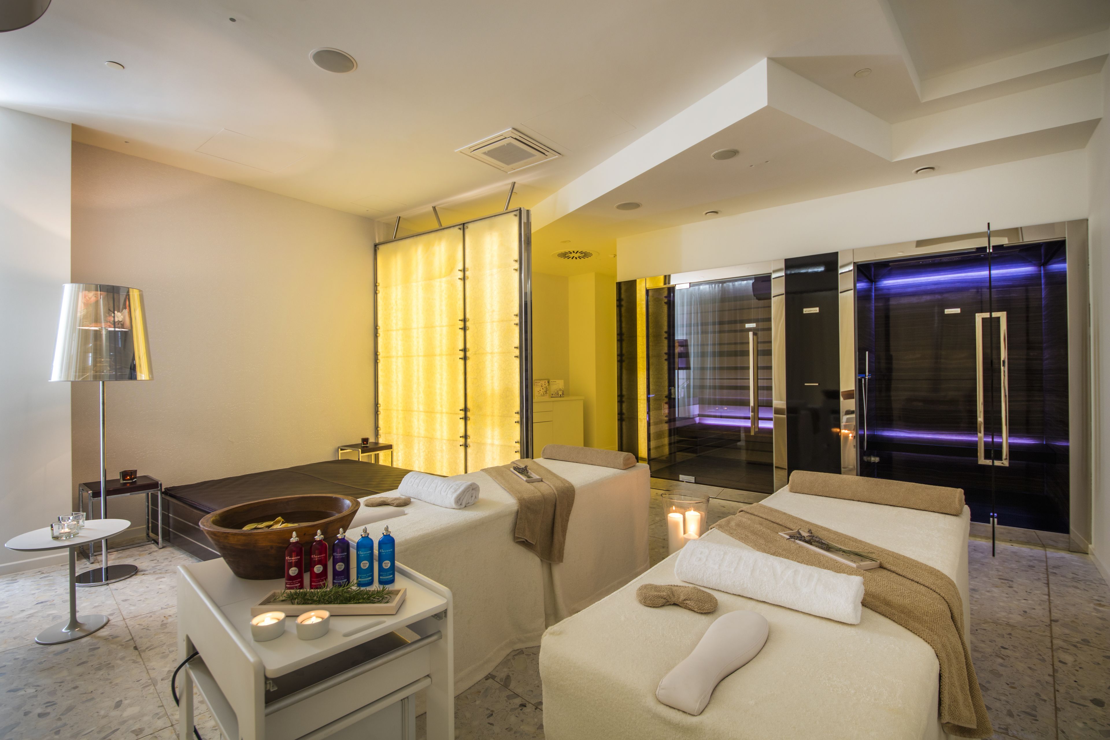 A modern hotel room with a large bed, a small bedside table with candles, a wooden bowl with fruits, and a massage table.