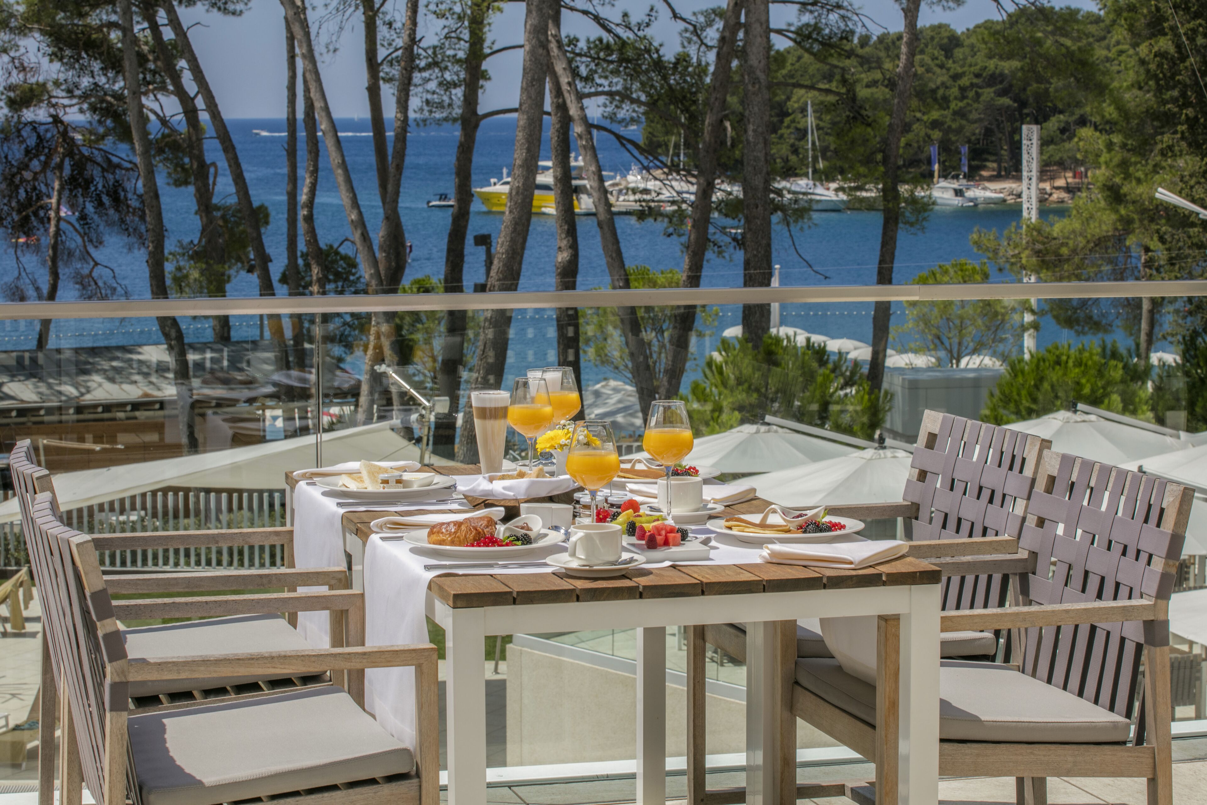 Une table à manger dressée avec des aliments et des boissons pour le petit-déjeuner, donnant sur une zone côtière avec des bateaux et des arbres.