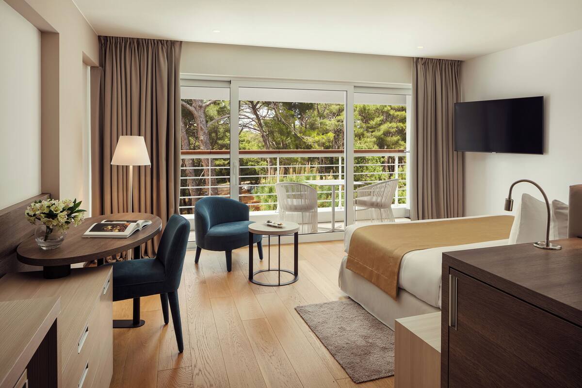 Ein Hotelzimmer mit Bett, Schreibtisch, Stuhl, Fernseher und einem Balkon mit Blick auf Bäume.