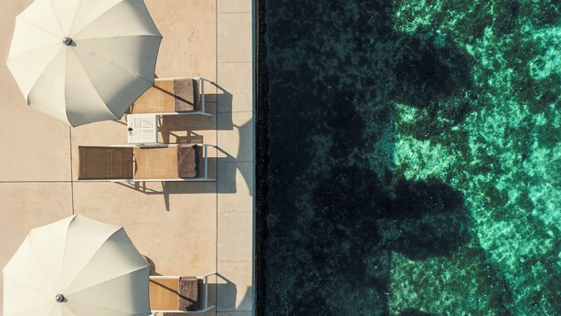 Vista aérea de un resort frente a la playa, con tumbonas y sombrillas, una piscina y agua turquesa cristalina.