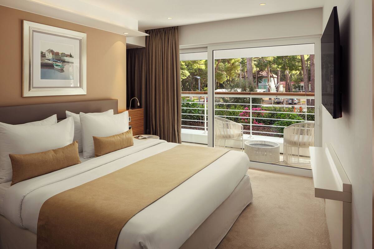 Ein Hotelzimmer mit einem großen Bett, einem Balkon mit weißen Stühlen und einem an der Wand befestigten Fernseher.