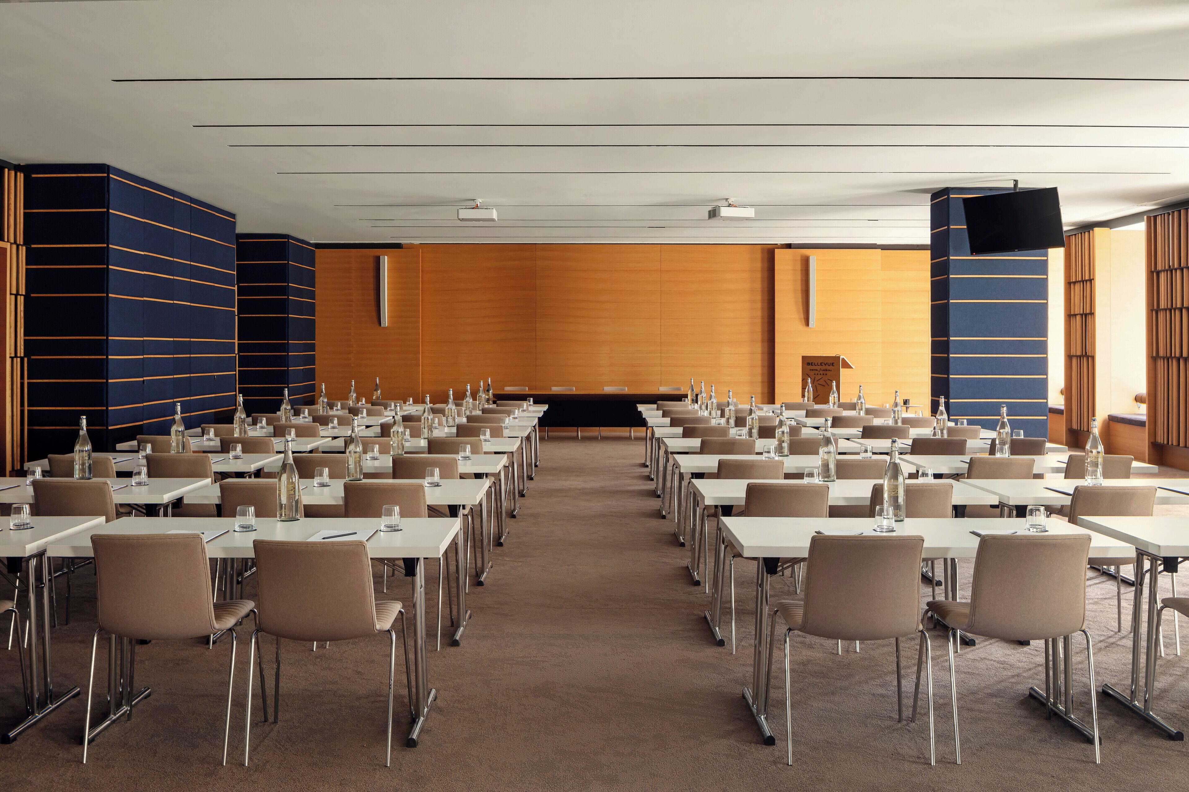 A conference room with rows of tables and chairs, a podium, and a large screen.
