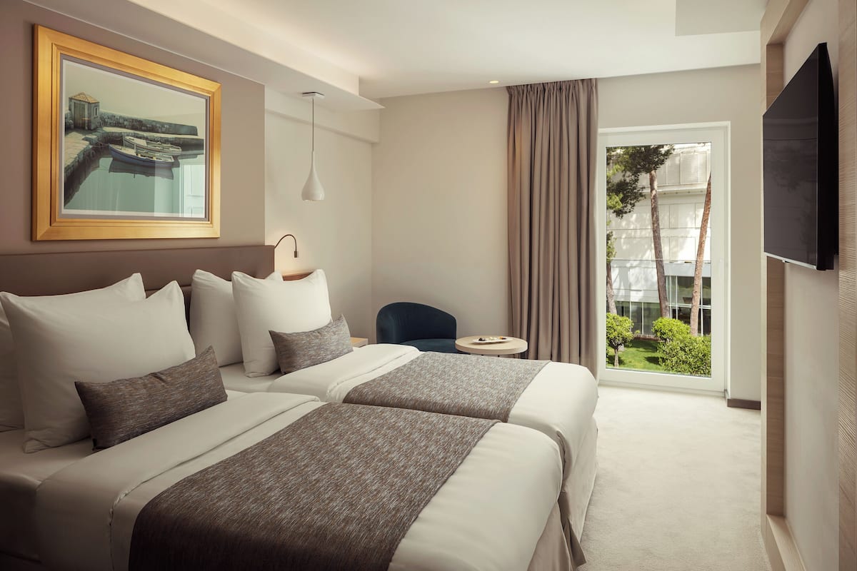 Ein Hotelzimmer mit zwei Betten, einem Fernseher, einem Gemälde und Blick ins Freie.