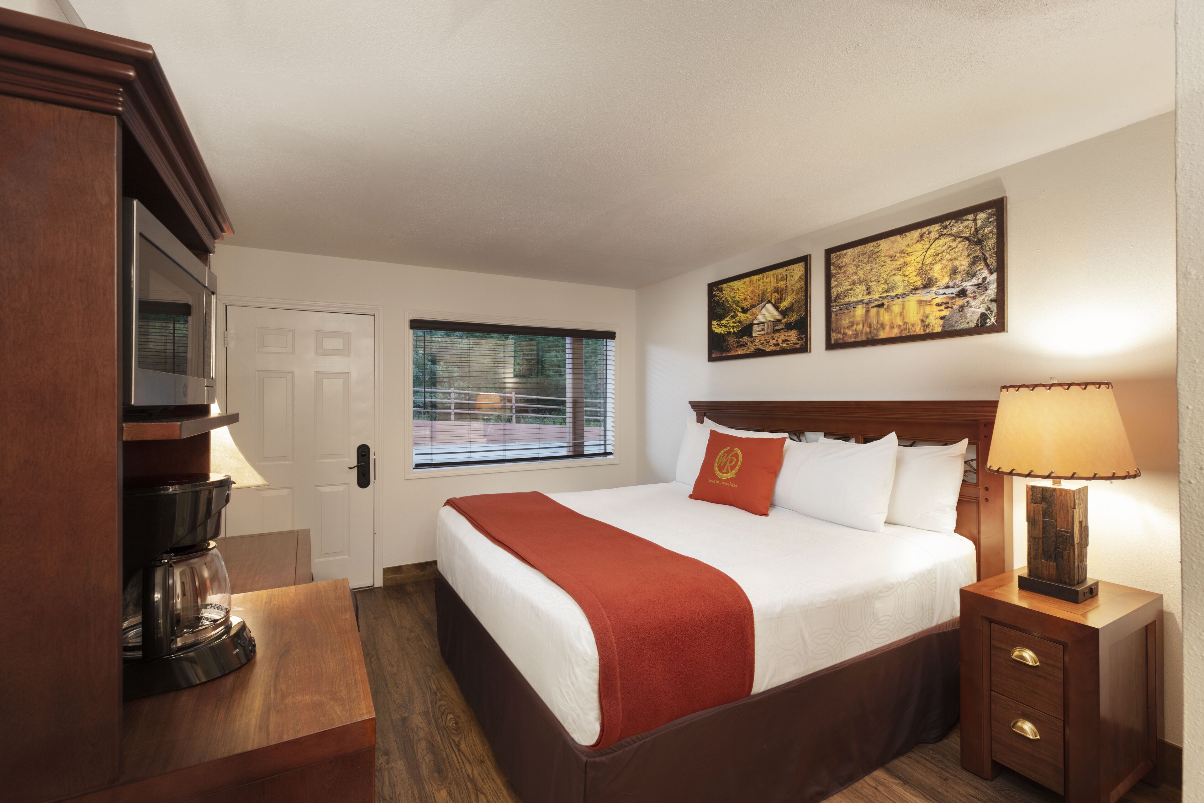 Habitación de hotel con cama, mesita de noche con lámpara, cafetera, ventana con persianas y dos cuadros en la pared.