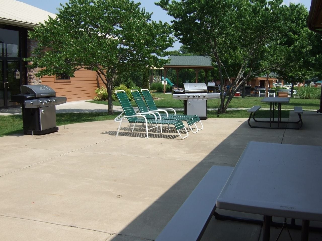 An outdoor patio with a grill, green lounge chairs, and picnic tables.