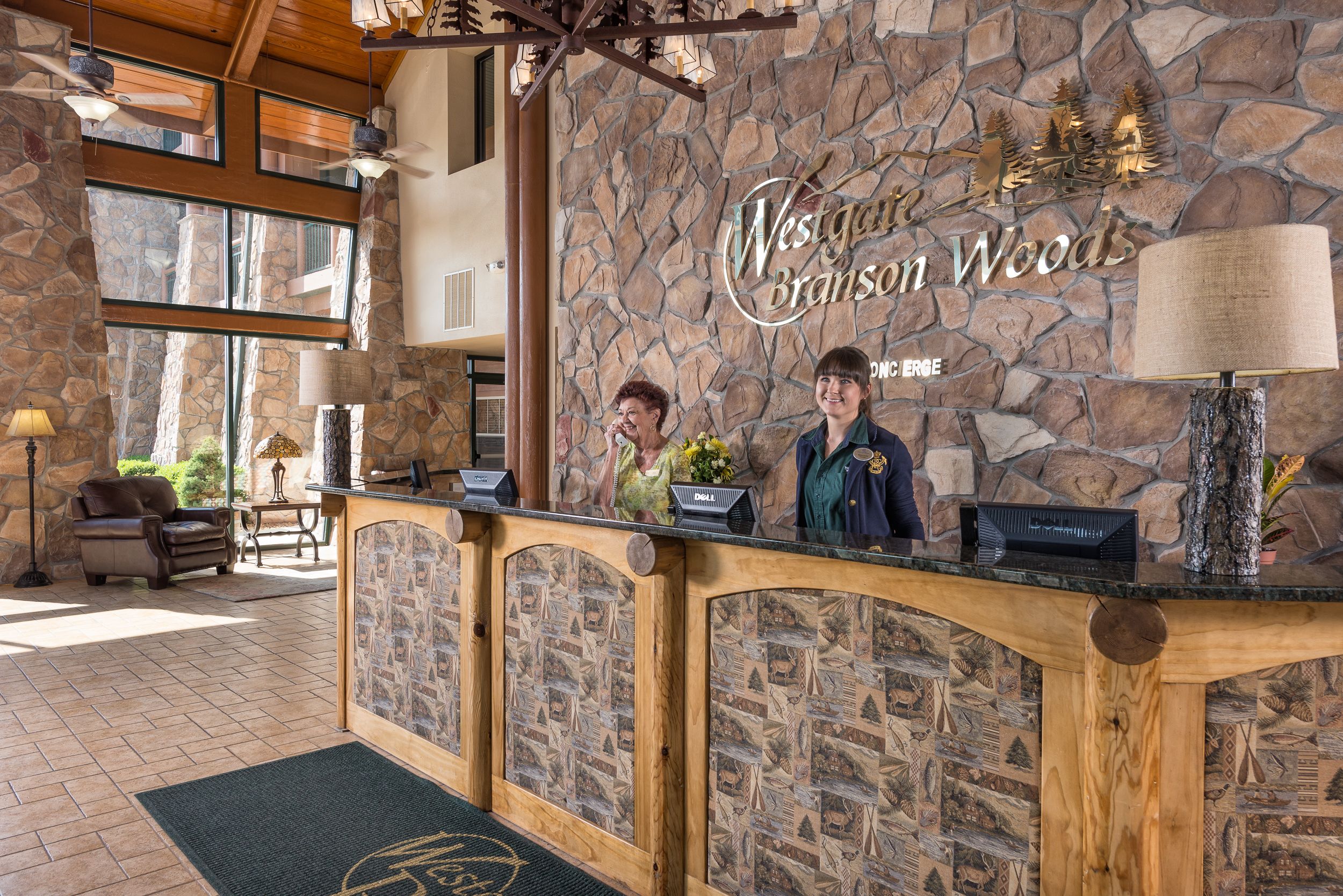 Un comptoir d’accueil à l’hôtel Westgate Branson Woods, avec un mur en pierre en arrière-plan, un comptoir en bois et une grande fenêtre laissant entrer la lumière naturelle.