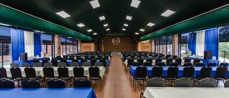 Une salle de conférence avec des rangées de tables et de chaises, une scène à l’avant et de grandes fenêtres de chaque côté.