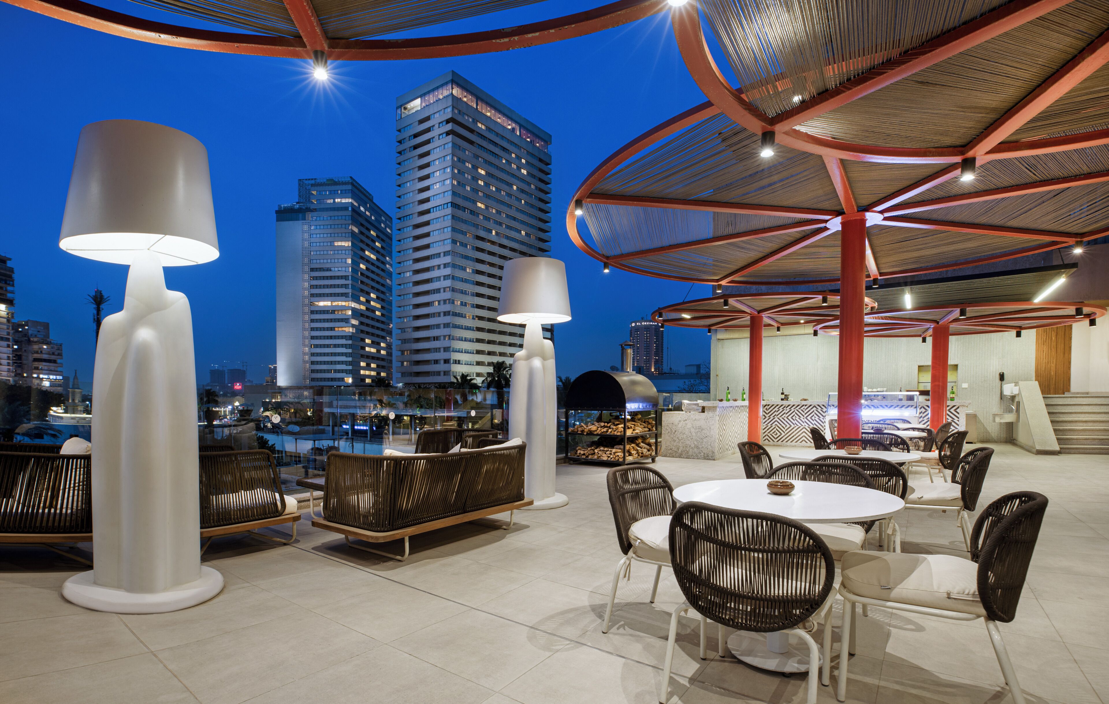 Een terras met ronde tafels en stoelen, een grote witte lamp en een uitzicht op hoge gebouwen bij zonsondergang.