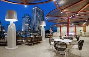 Un espacio para comer al aire libre con mesas y sillas redondas, una lámpara blanca grande y vistas de edificios altos al anochecer.