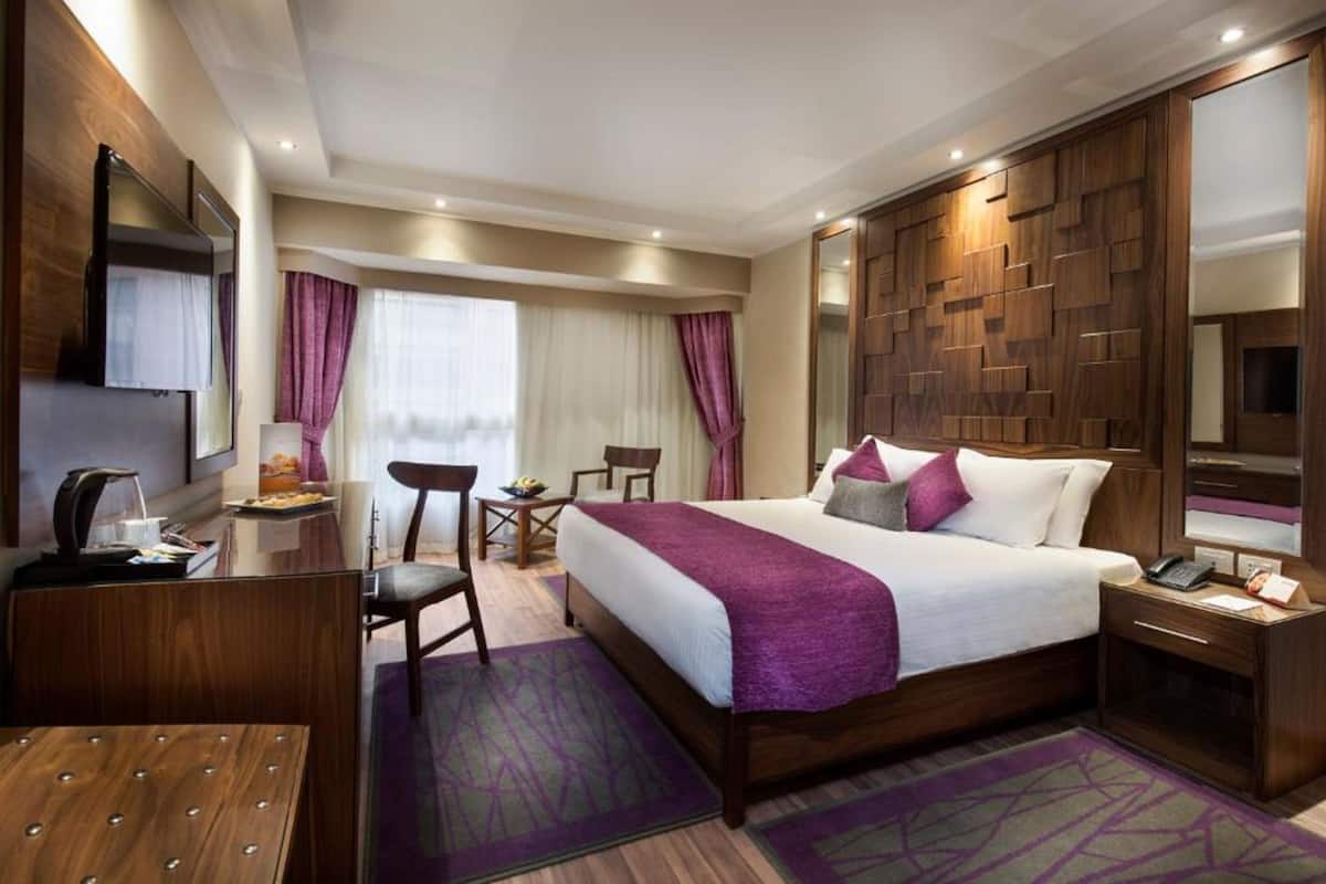 A hotel room with a large bed, a desk with a telephone, a chair, a small table with a lamp, and a TV mounted on the wall.