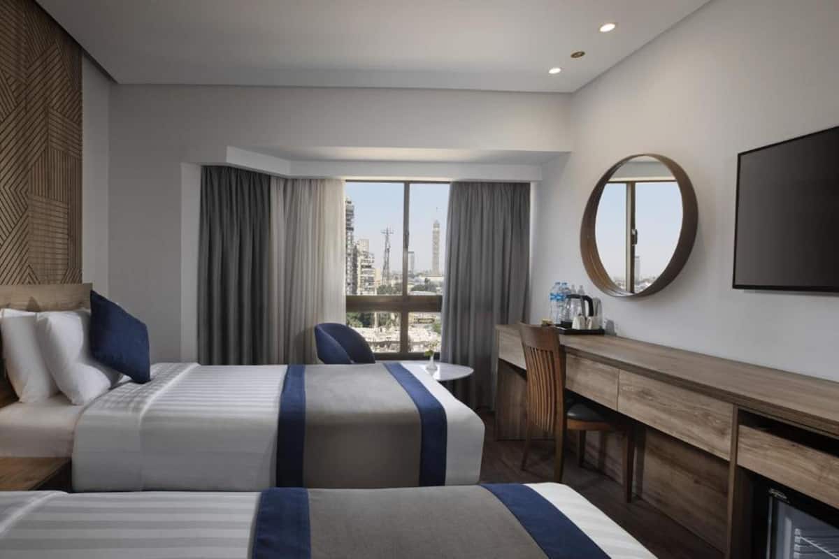 A hotel room with two beds, a desk, a chair, a round mirror, and a TV.