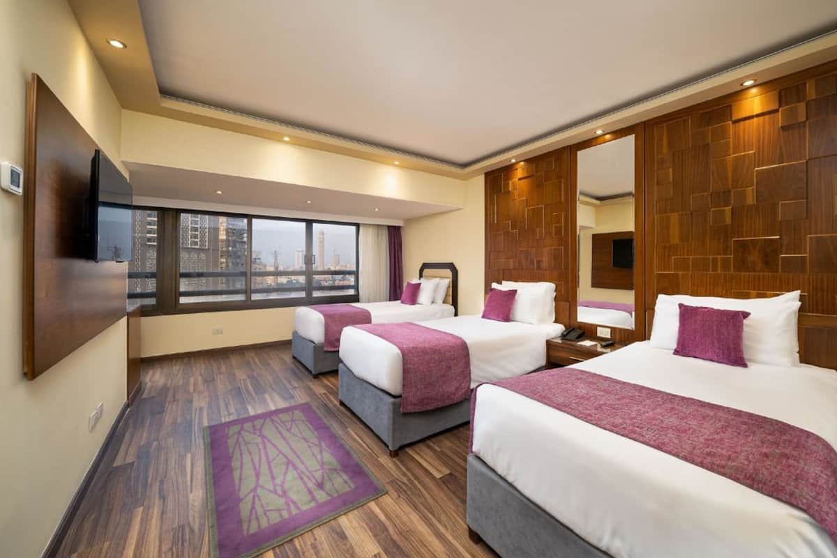 A hotel room with three beds, a large window with a city view, a wooden headboard, and a flat-screen TV mounted on the wall.