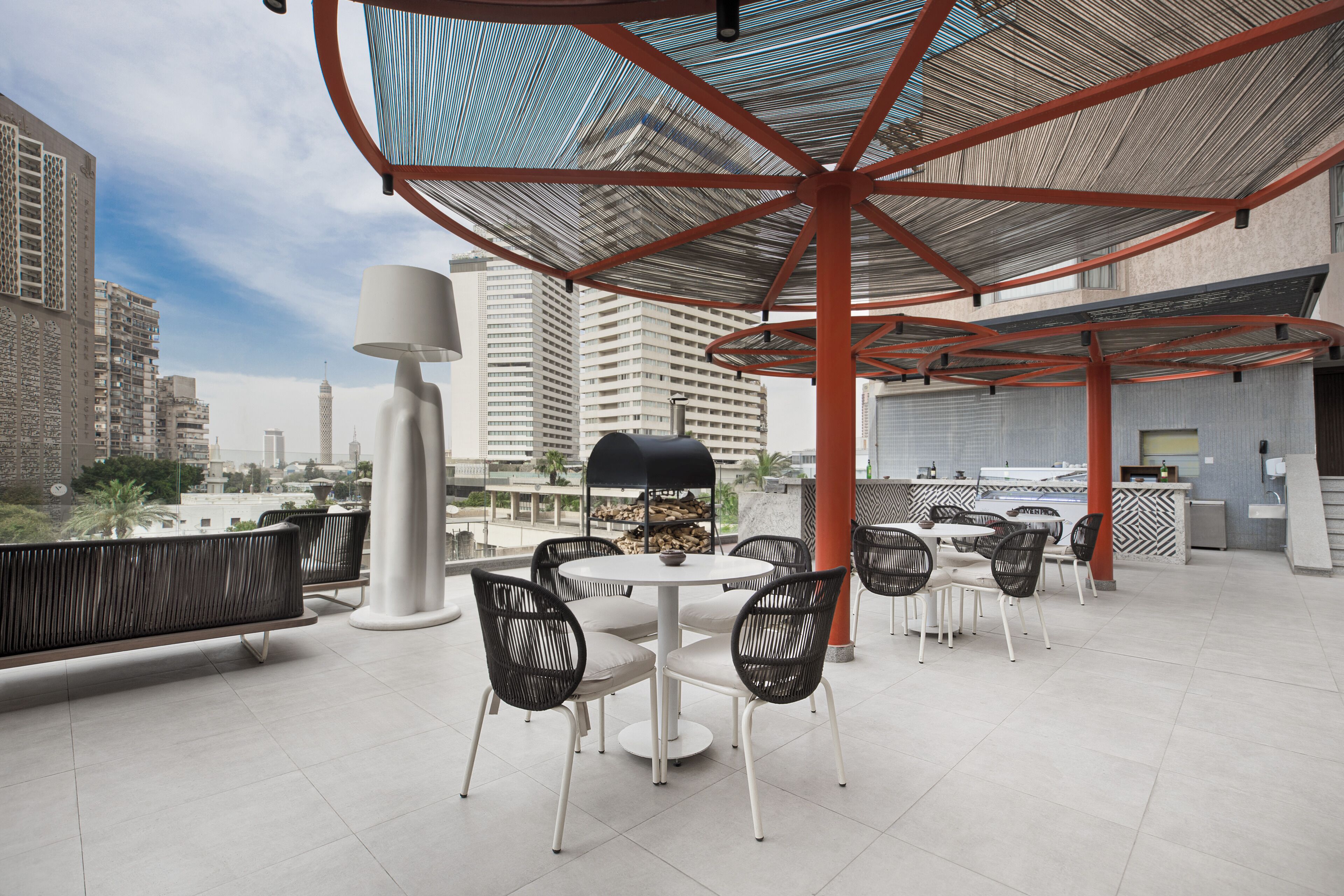 An outdoor seating area with tables and chairs, a large umbrella structure, and a view of city buildings in the background.